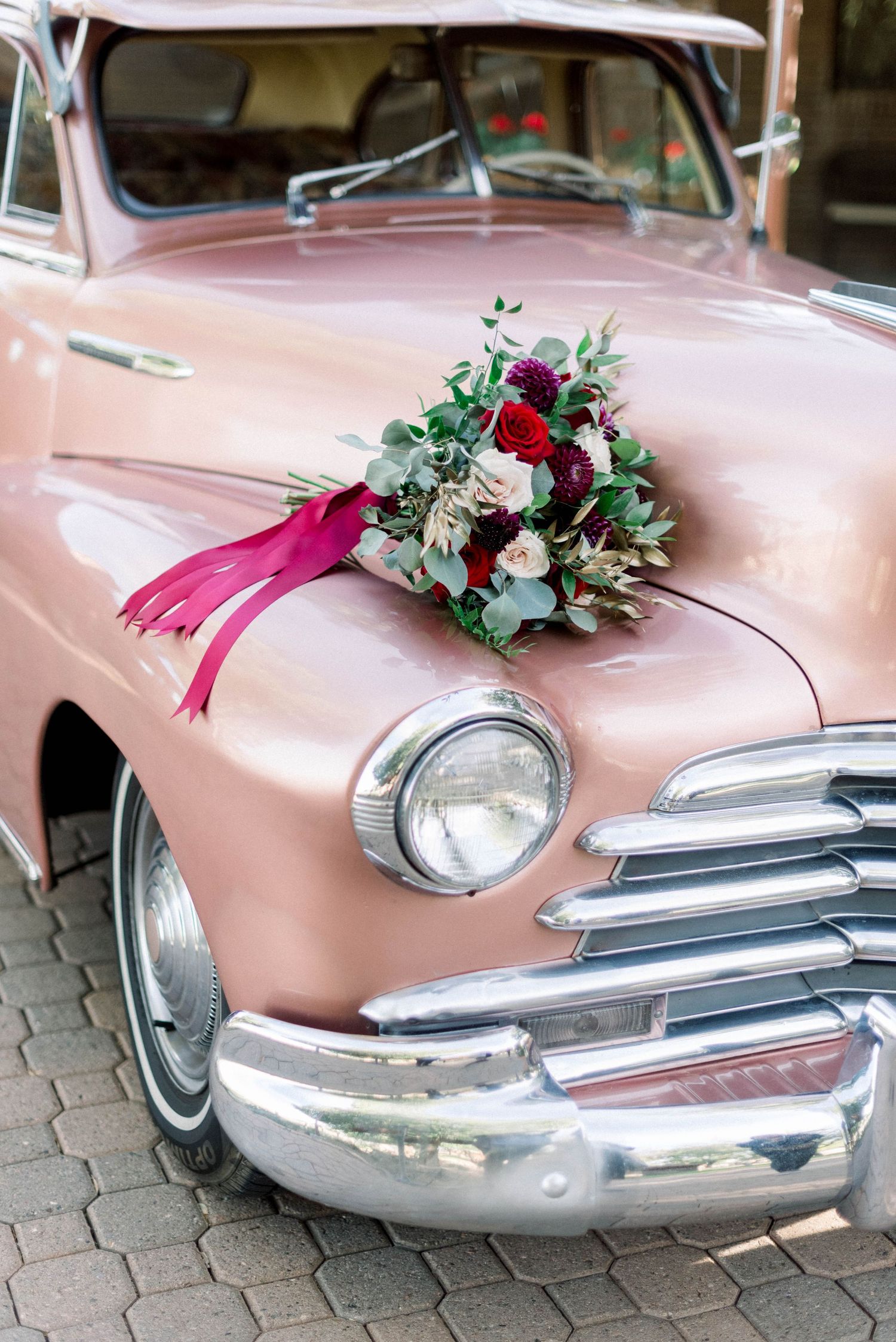 Classic pink vintage car decorated with burgundy and white floral arrangement on hood.