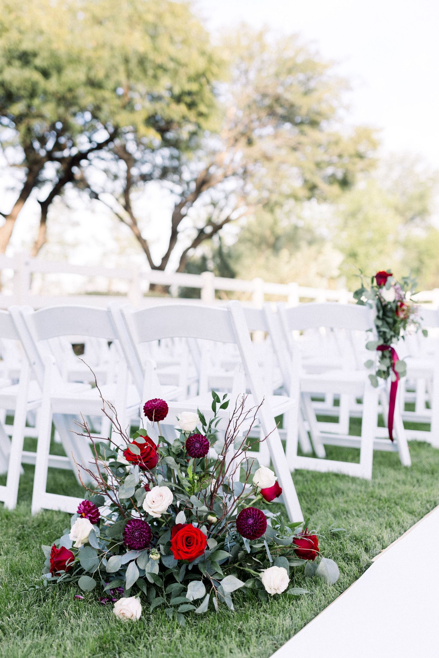 Beautiful red and white floral arrangements line an outdoor wedding aisle with white folding chairs.