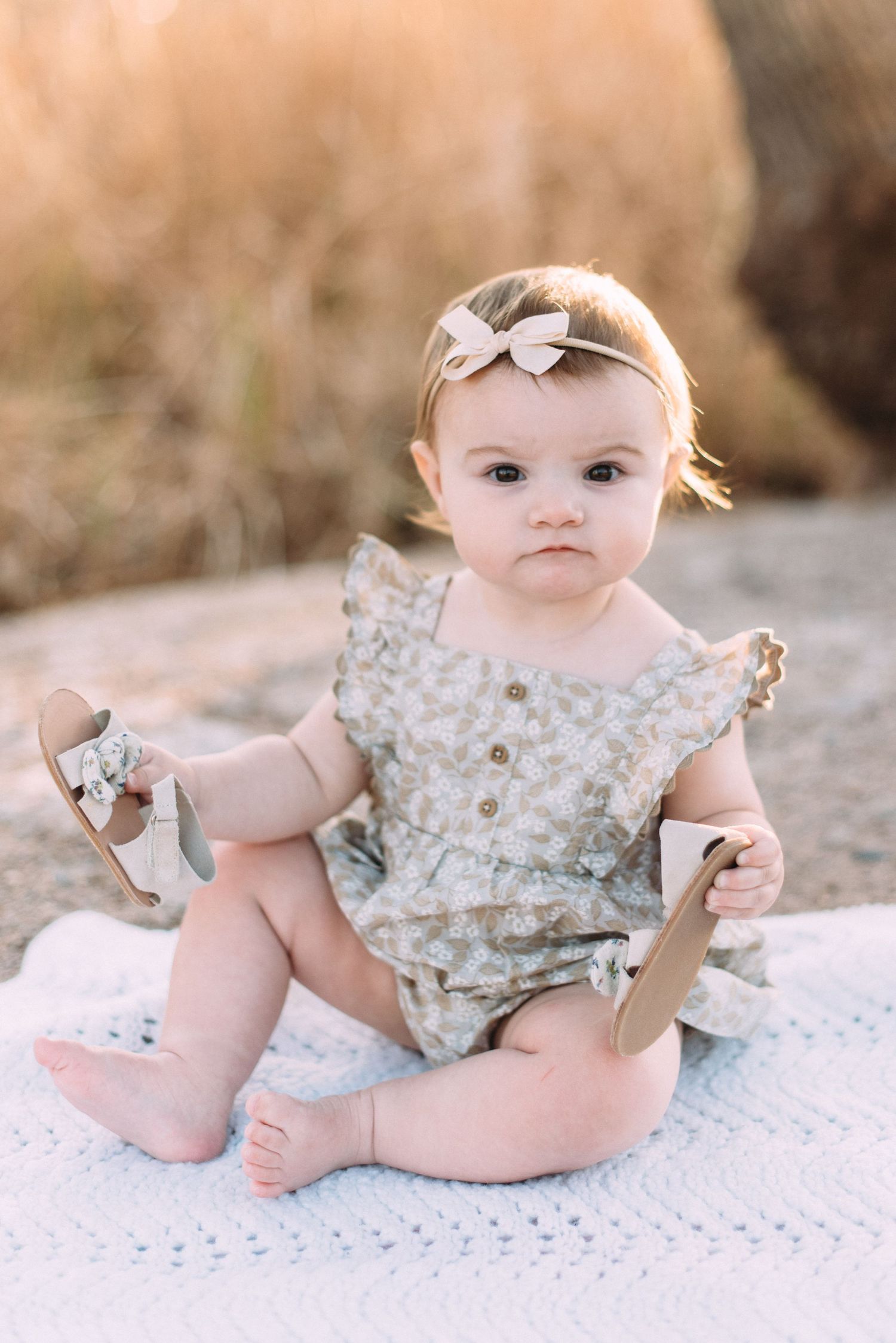 Baby in floral romper with bow headband sitting on blanket during outdoor photoshoot.
