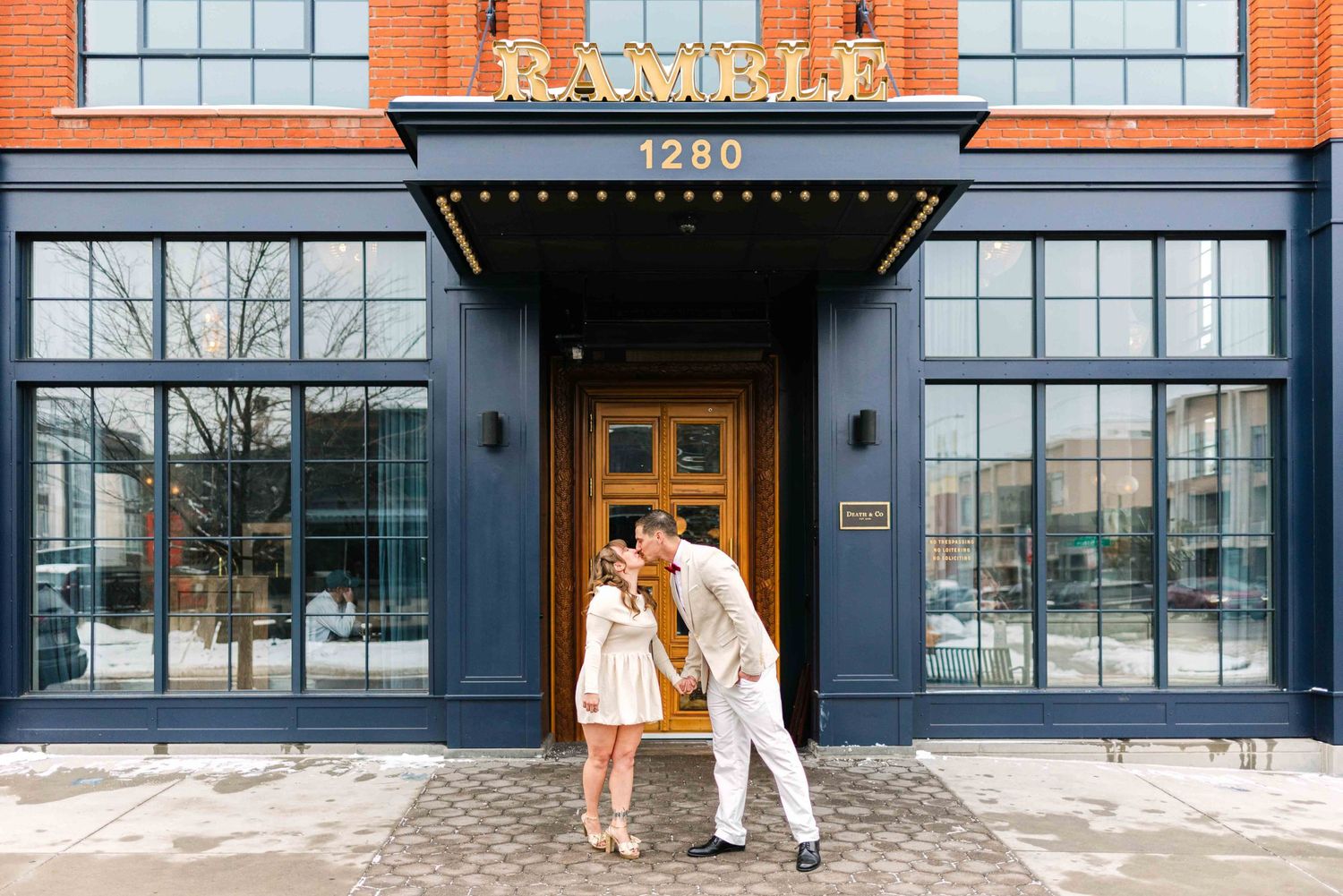 A couple poses in front of a brick building with large windows and an elegant black awning entrance.