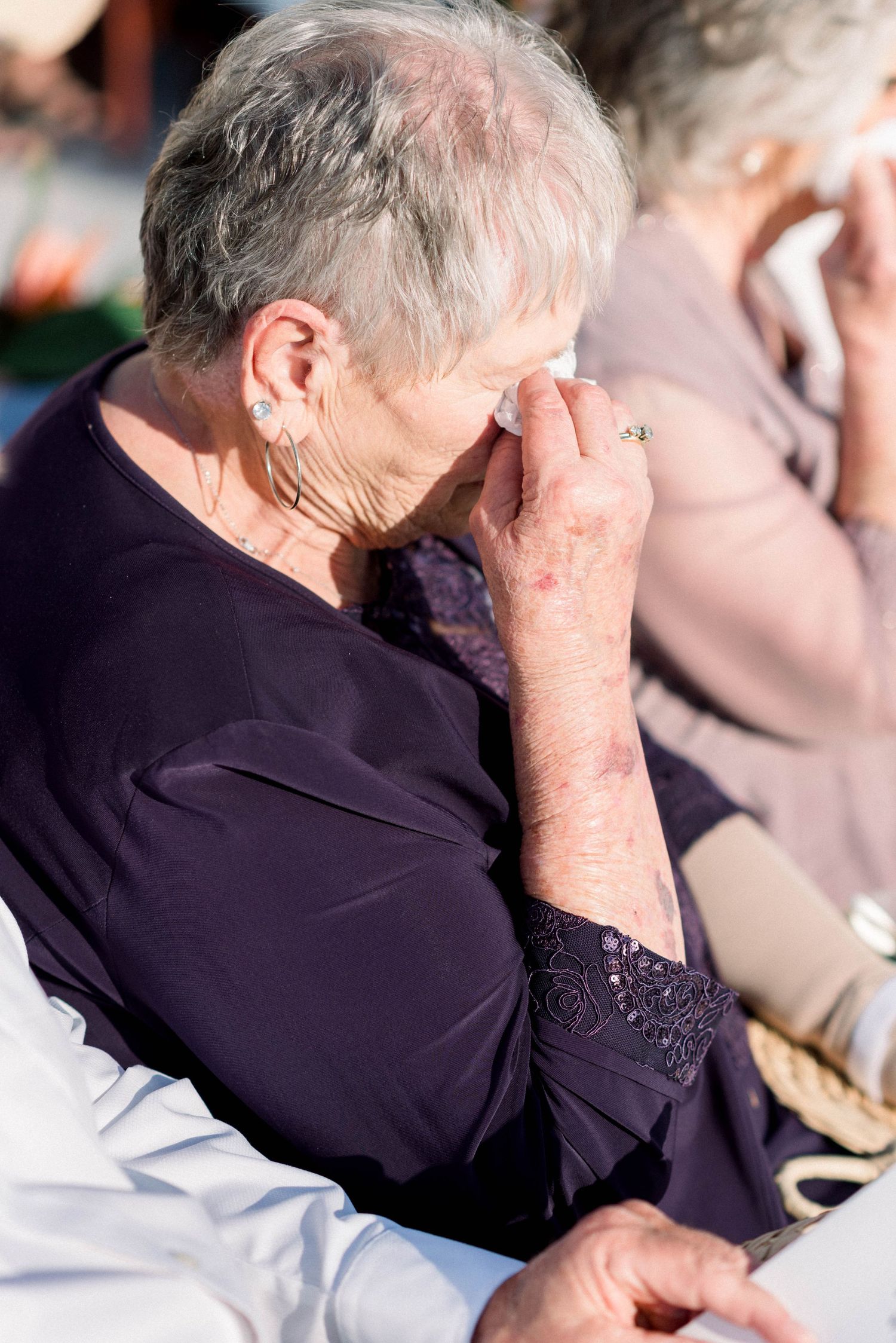 An elderly person in a purple blouse holding their hand to their face during an emotional moment at an event.