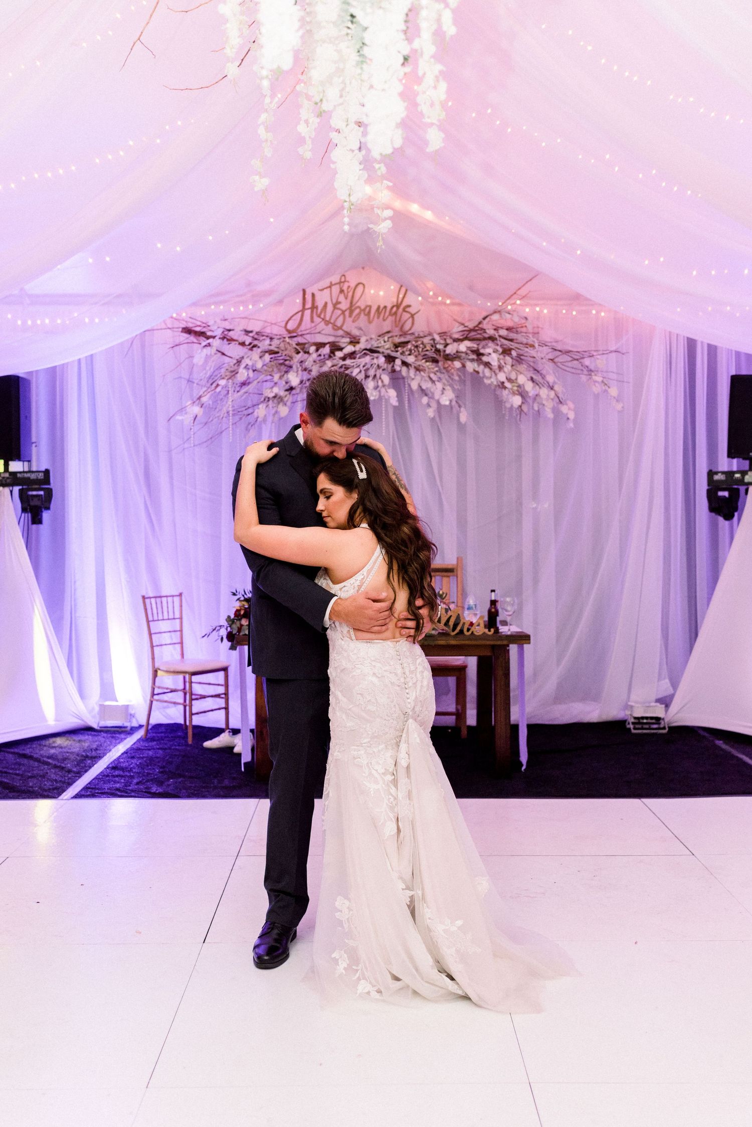 Romantic dance moment under floral chandelier at purple-lit wedding reception.