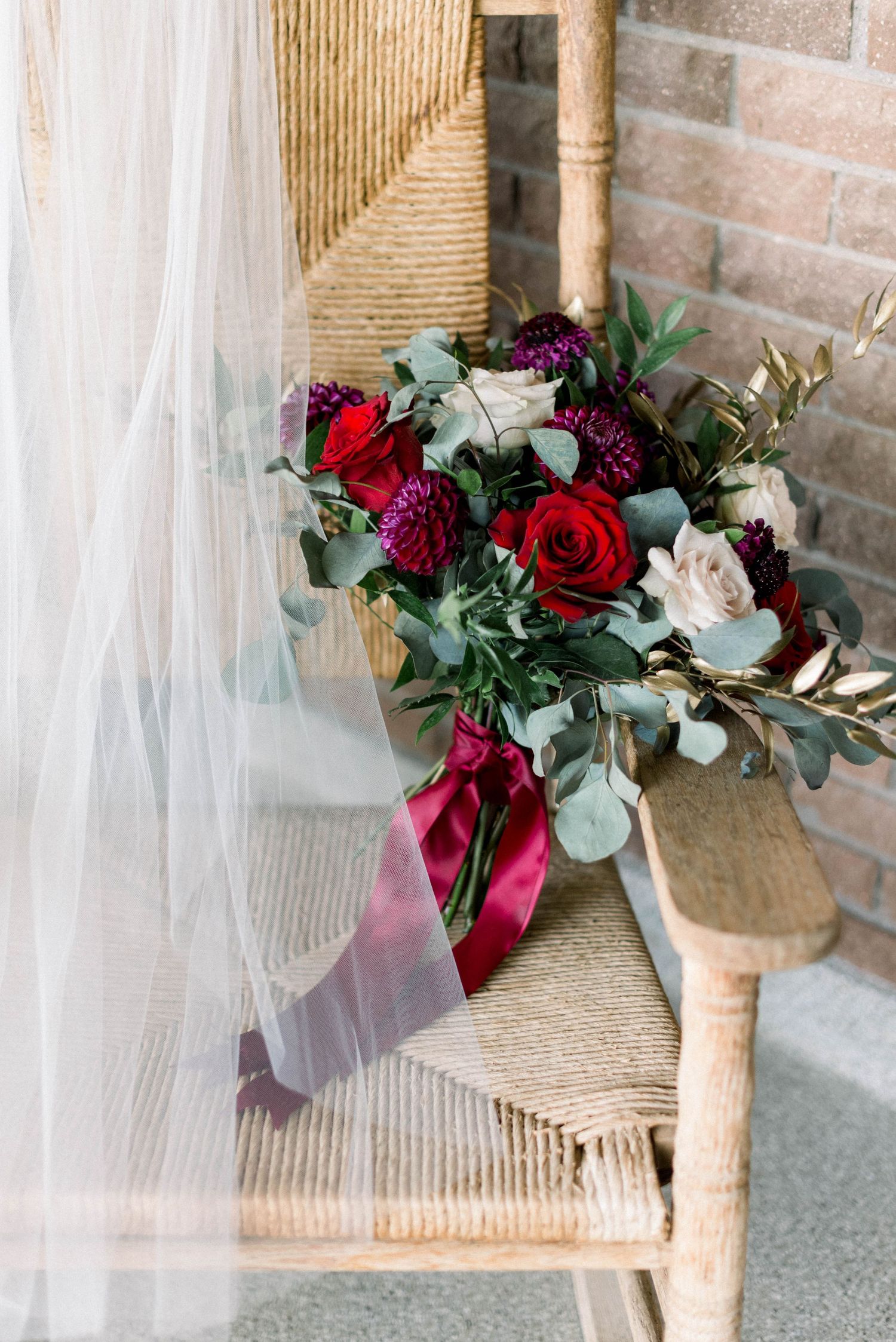 Romantic bouquet with red roses and burgundy flowers tied with velvet ribbon rests on wicker chair.
