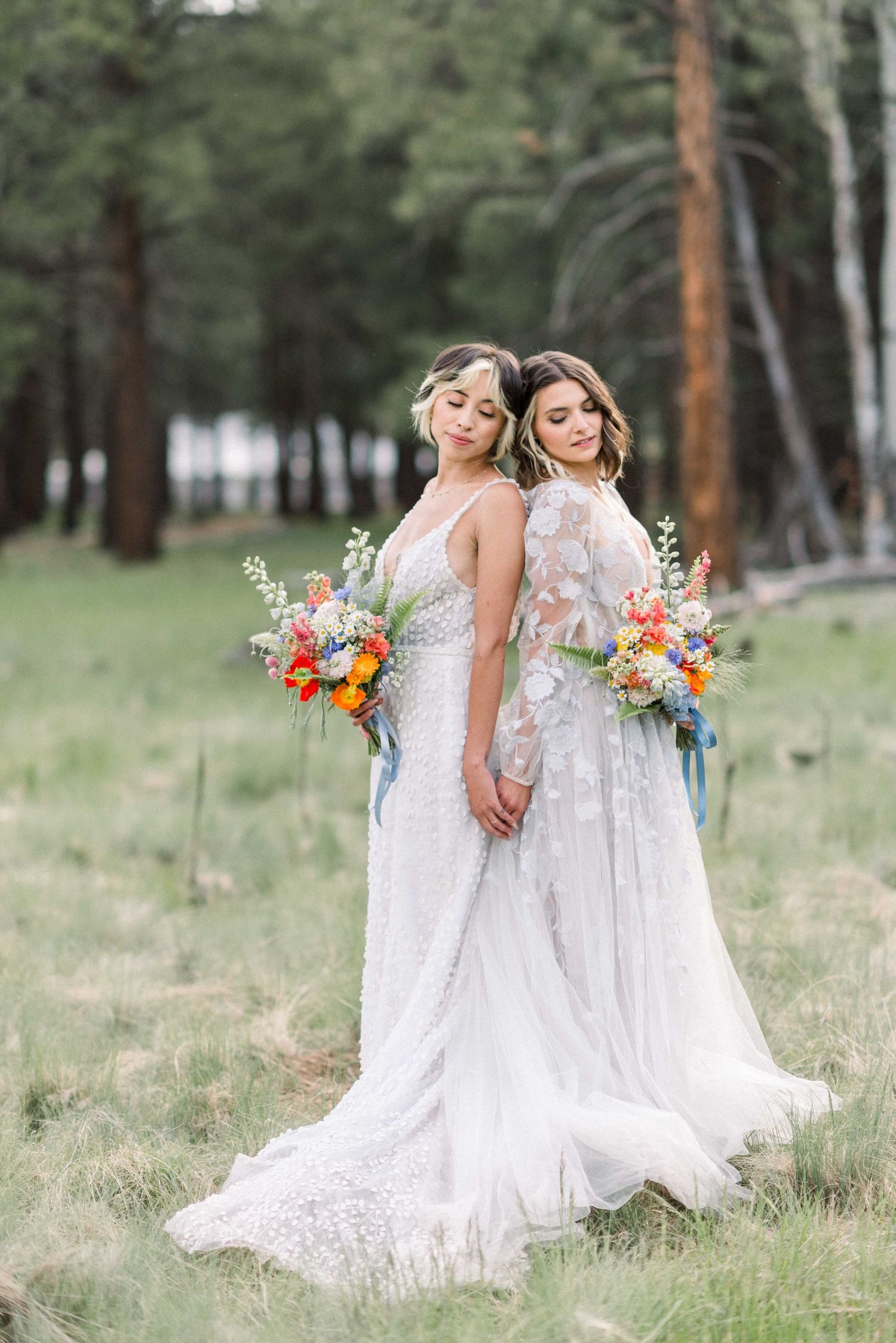 Two brides in elegant white gowns pose back-to-back holding colorful bouquets in a pine forest.