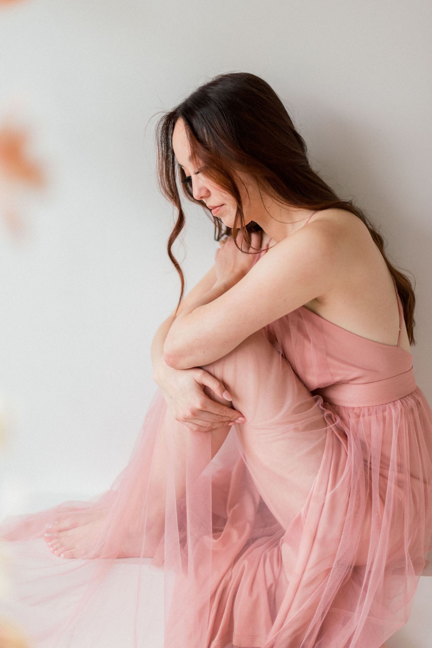 A person in a flowing pink tulle dress poses gracefully against a white wall in a soft, romantic portrait.