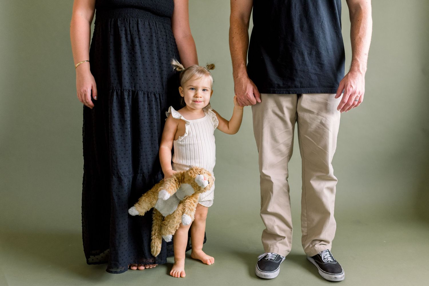 Family standing together with toddler holding stuffed animal against minimal studio background.