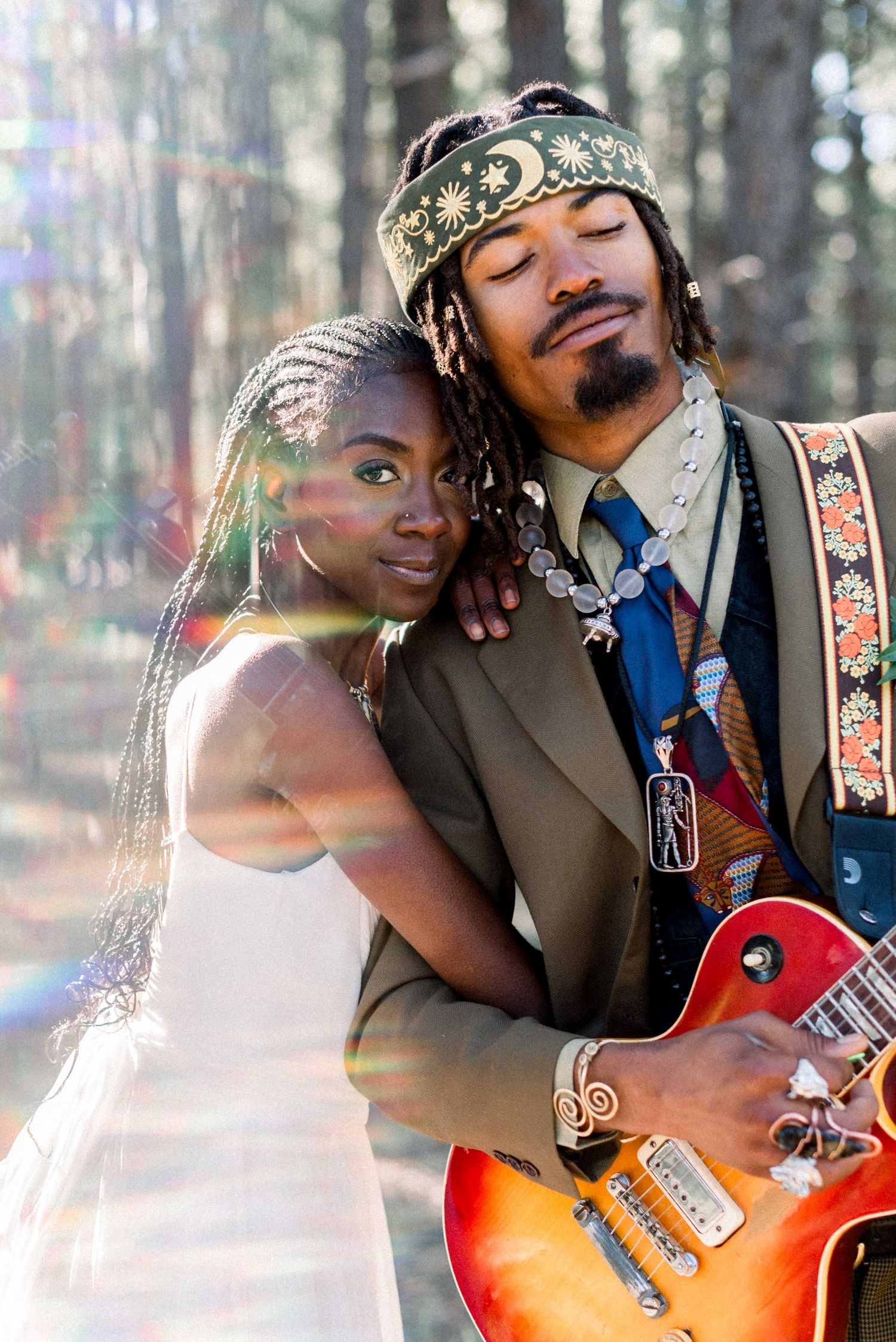 A sun-drenched wedding portrait of a couple embracing while the groom holds his guitar in a forest setting.