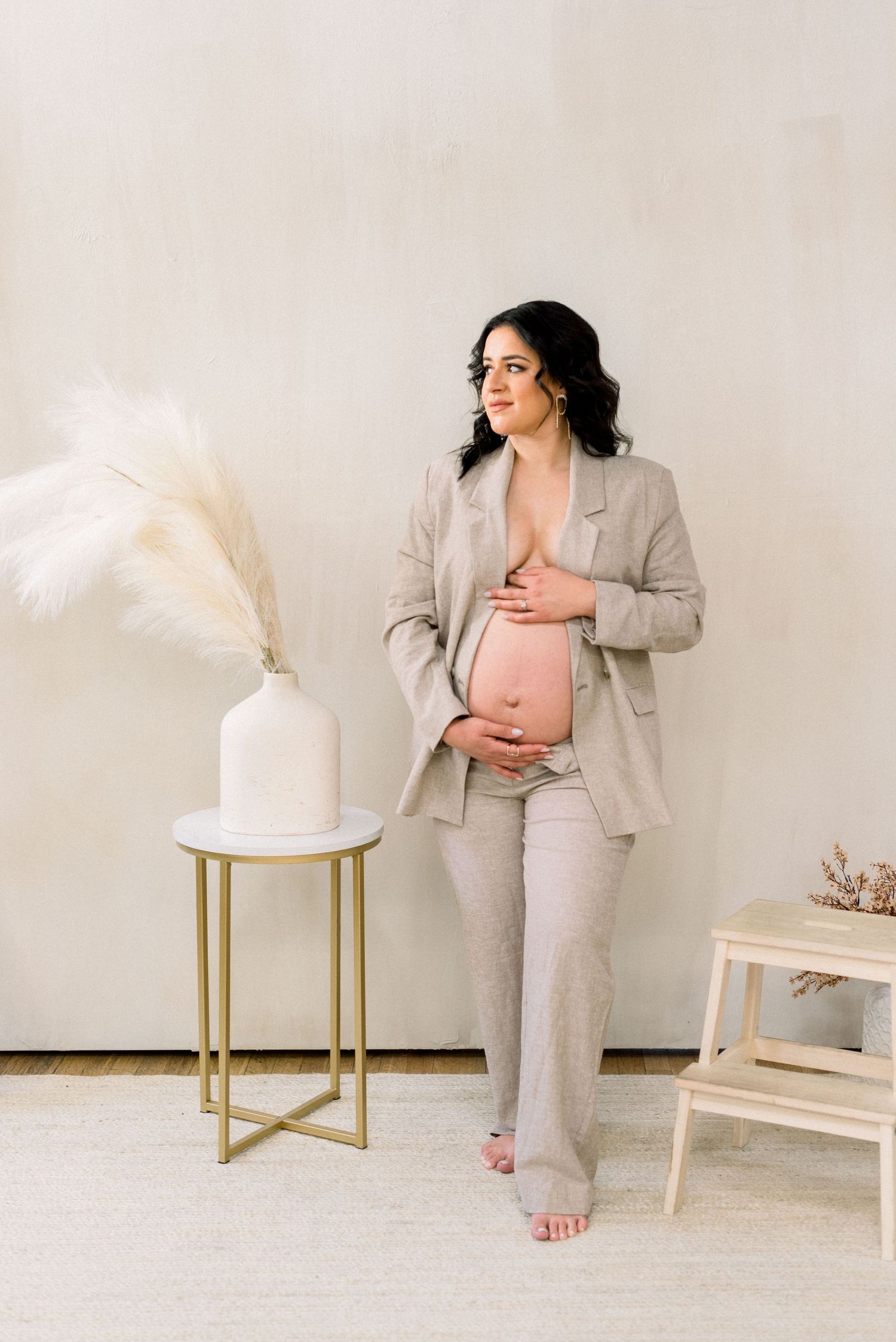 Pregnant woman in grey suit poses beside decorative vase with pampas grass.