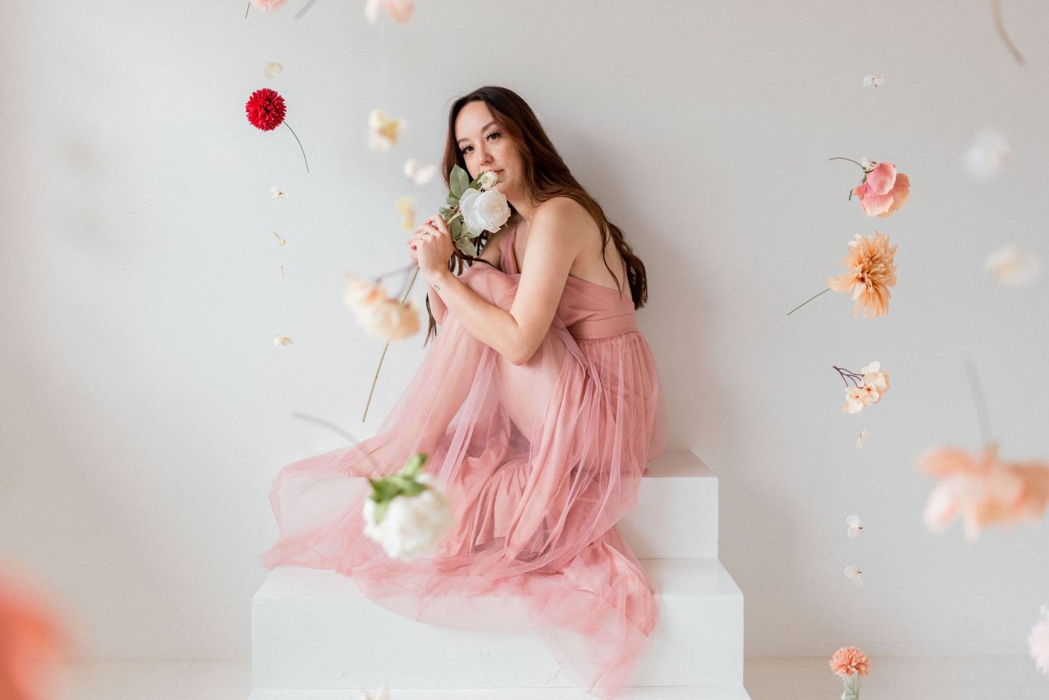 Dreamy portrait on white steps with flowing pink gown and floating flower petals.