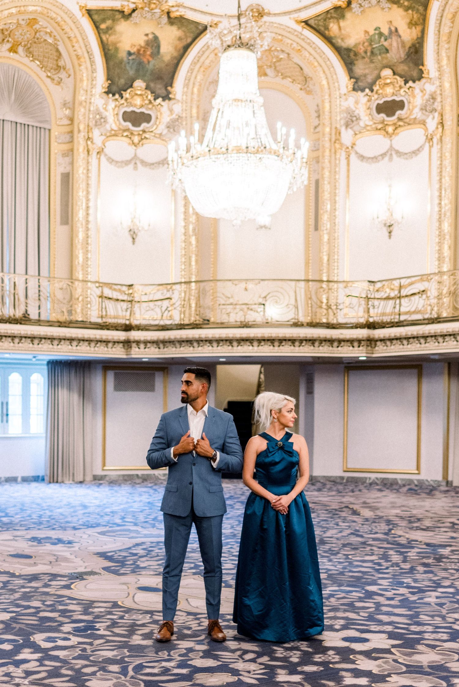 Elegant couple poses in opulent ballroom with crystal chandelier and ornate decor.