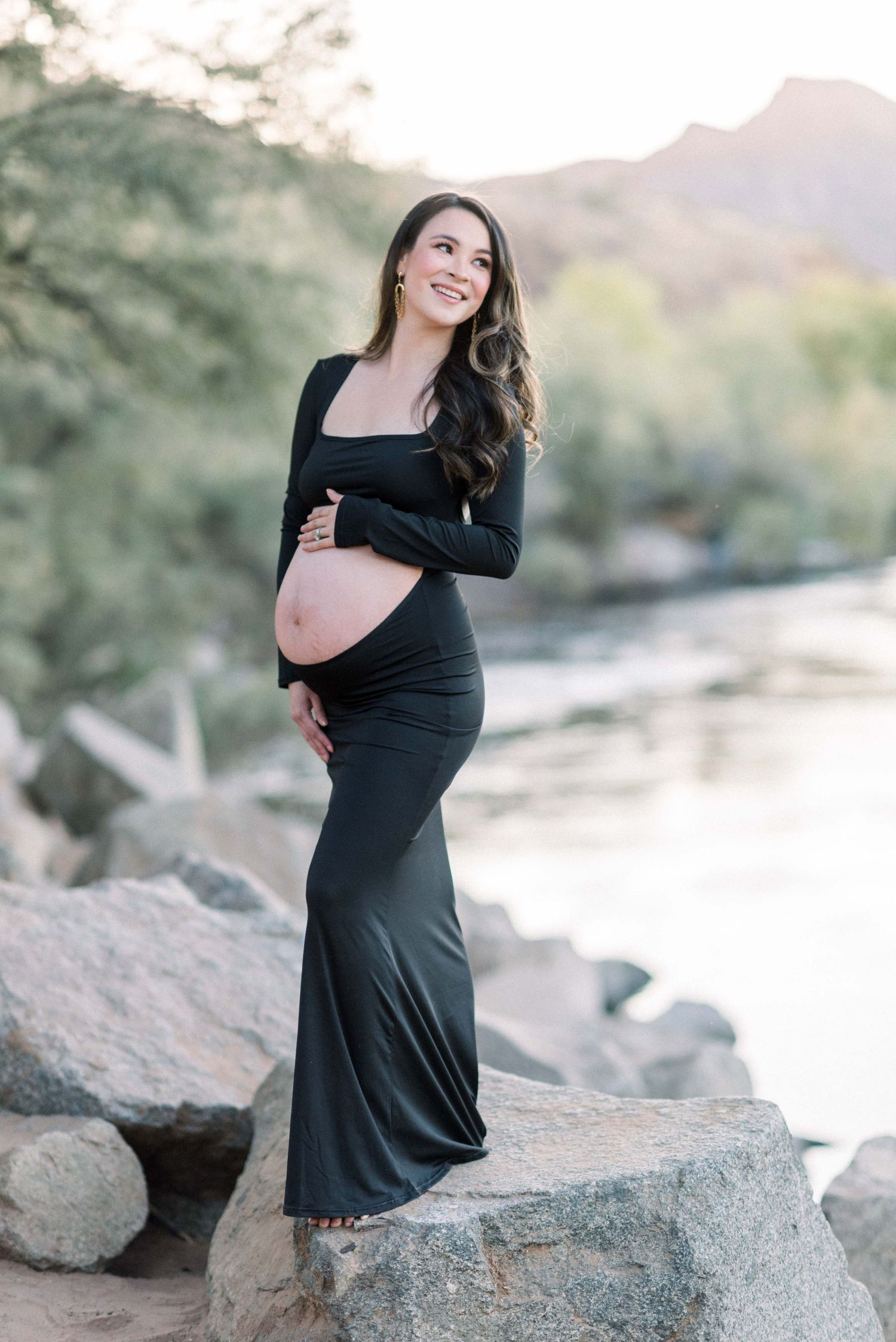 Maternity portrait in black dress standing on rocks by mountain river.