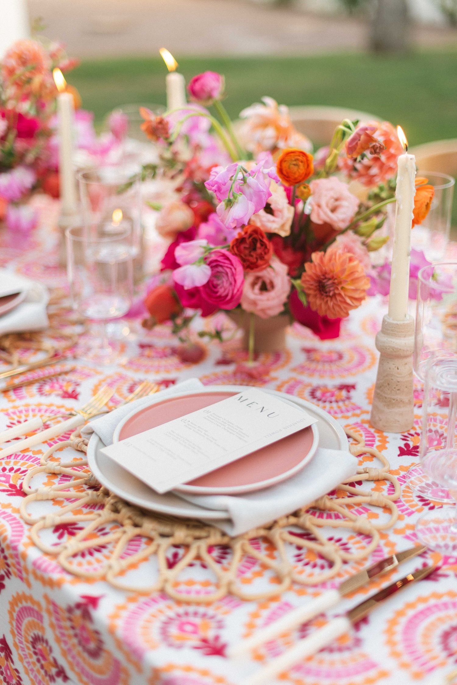 Close-up of wedding table decor with coral and pink flowers, white candles, and patterned linens.