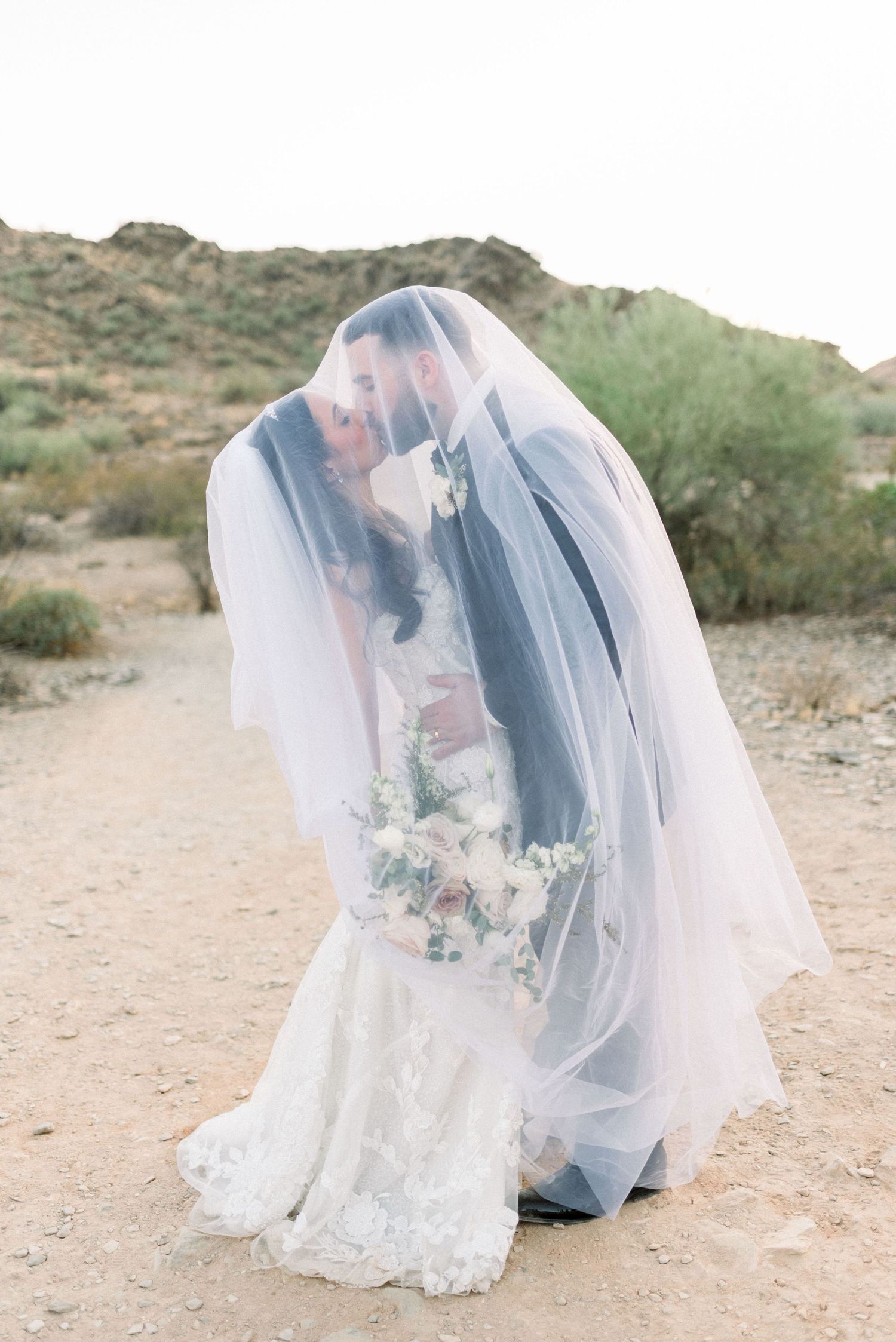 A romantic wedding photo series showing a couple under a flowing white veil in a desert landscape.