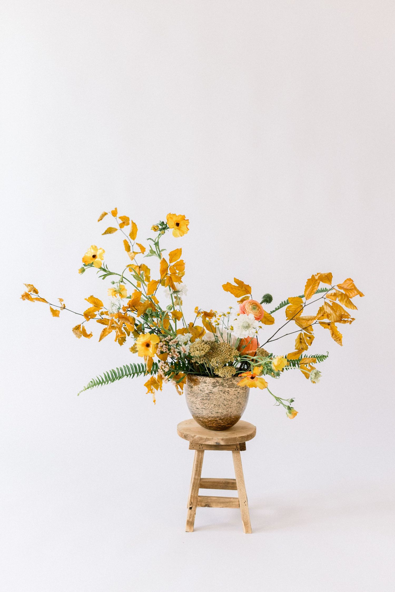 Yellow and orange floral arrangement in a rustic pot displayed on a wooden stool against white background.