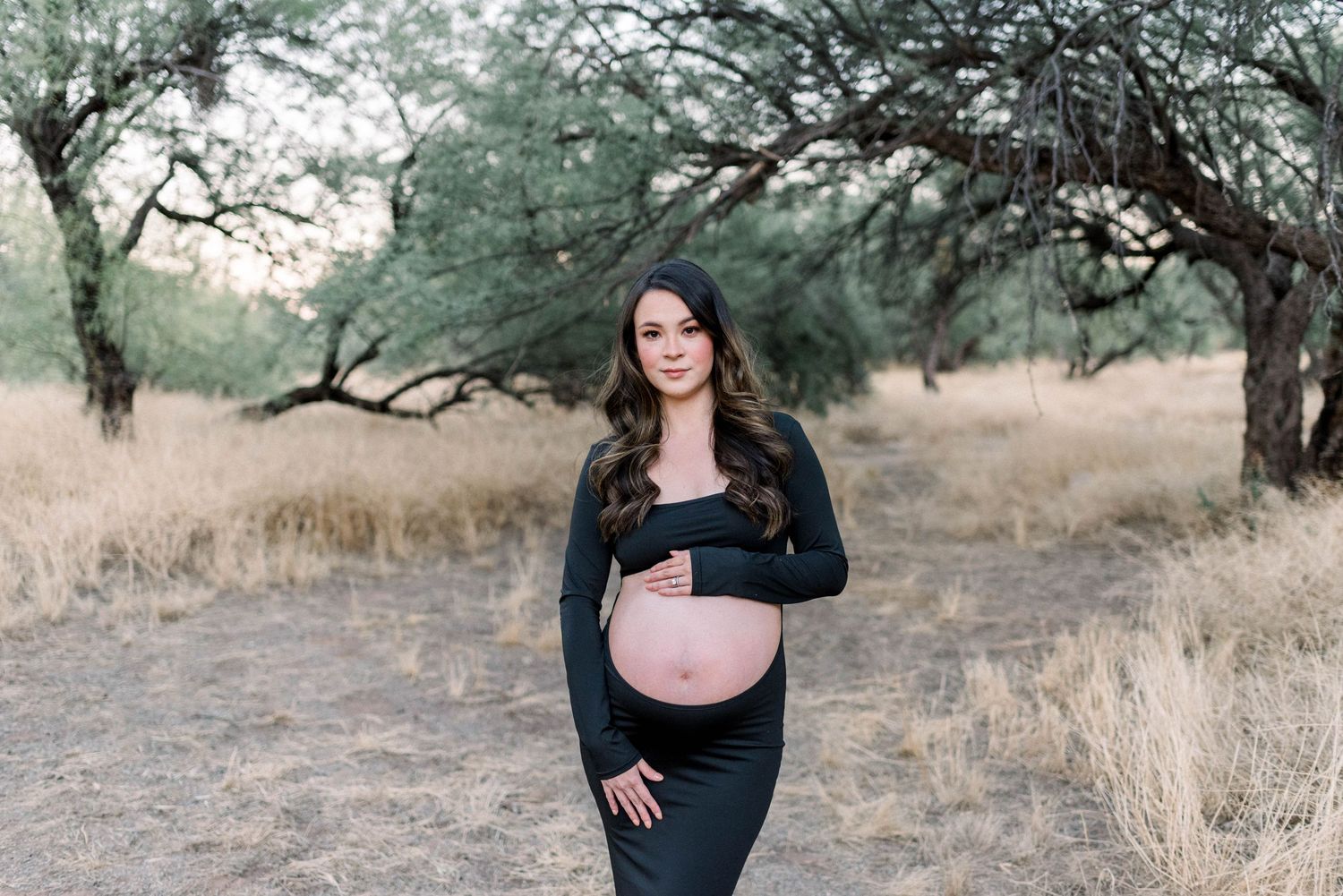 A woman in a black sweater poses outdoors in a rustic field setting with tall grass and trees at sunset.