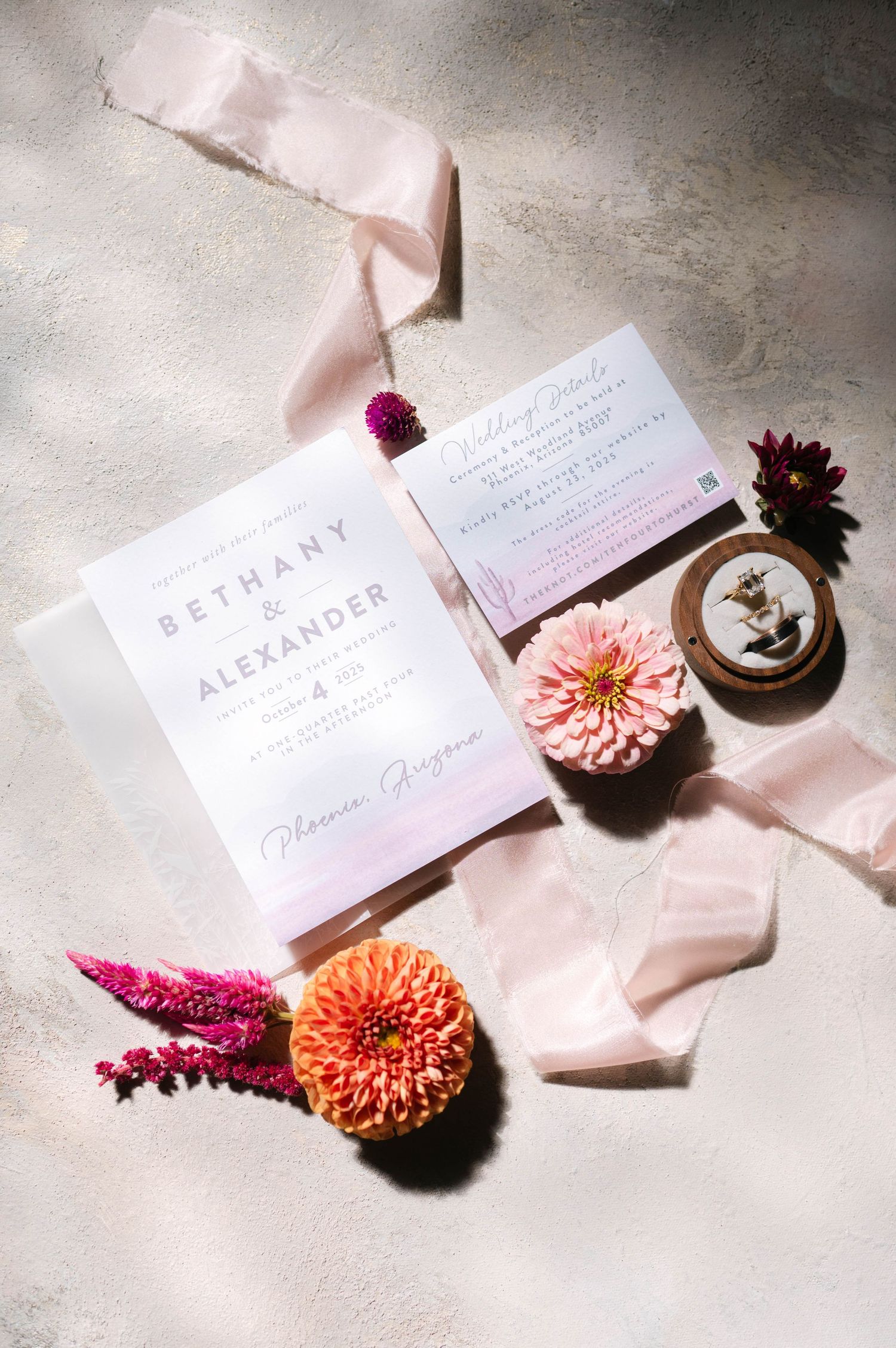 Wedding invitation suite styled with pink silk ribbons and vibrant flowers on stone background.