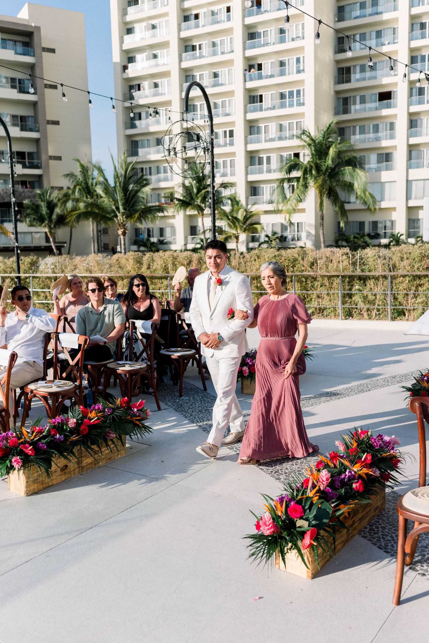 Wedding ceremony processional at a beachfront venue with tropical palm trees and high-rise buildings.