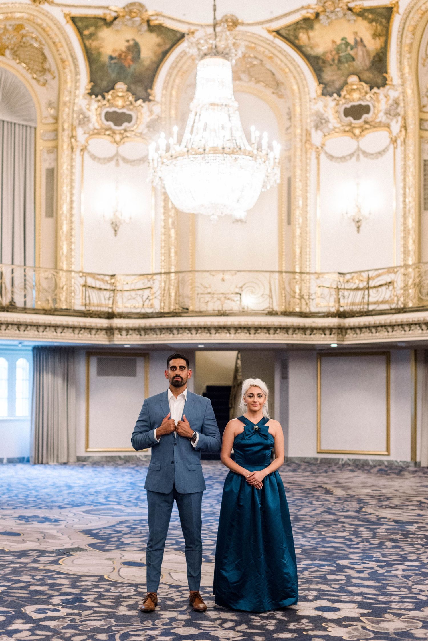 Well-dressed pair stands in luxurious ballroom with baroque architectural details.