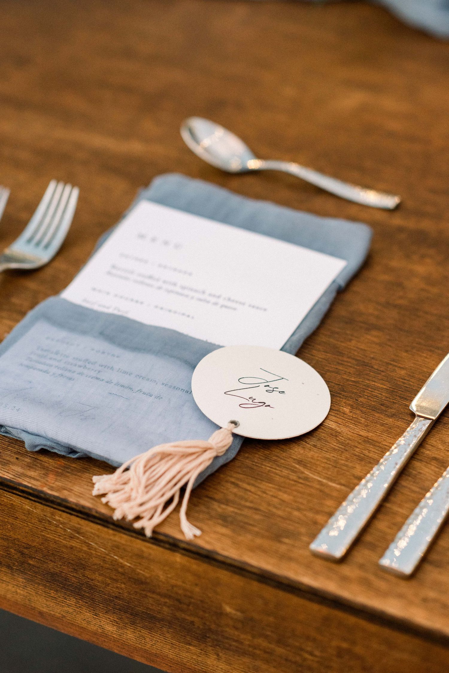 Elegant place setting with gray napkin and round monogrammed tag with tassel on wooden table.