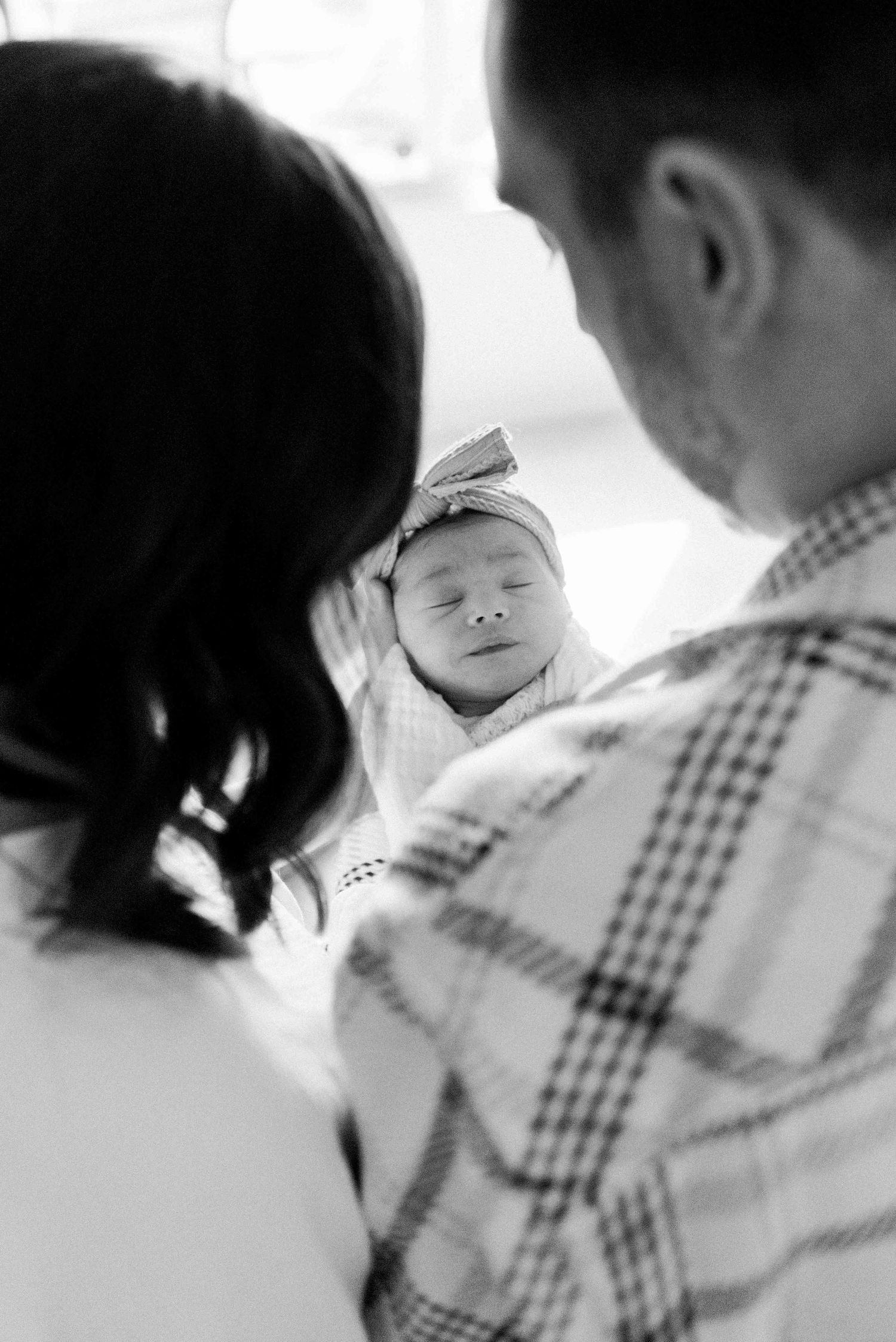 A peaceful sleeping newborn with a bow headband is held between two loving parents.