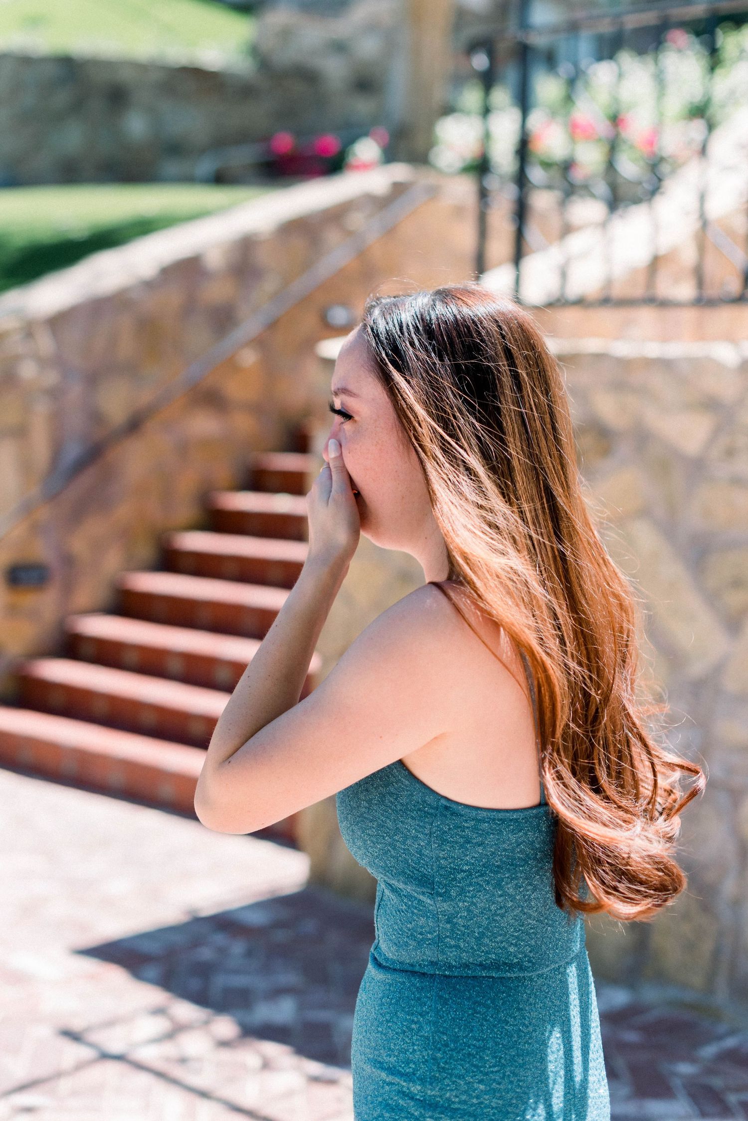 Woman in teal dress reacts emotionally on outdoor stone staircase with sunlit background.