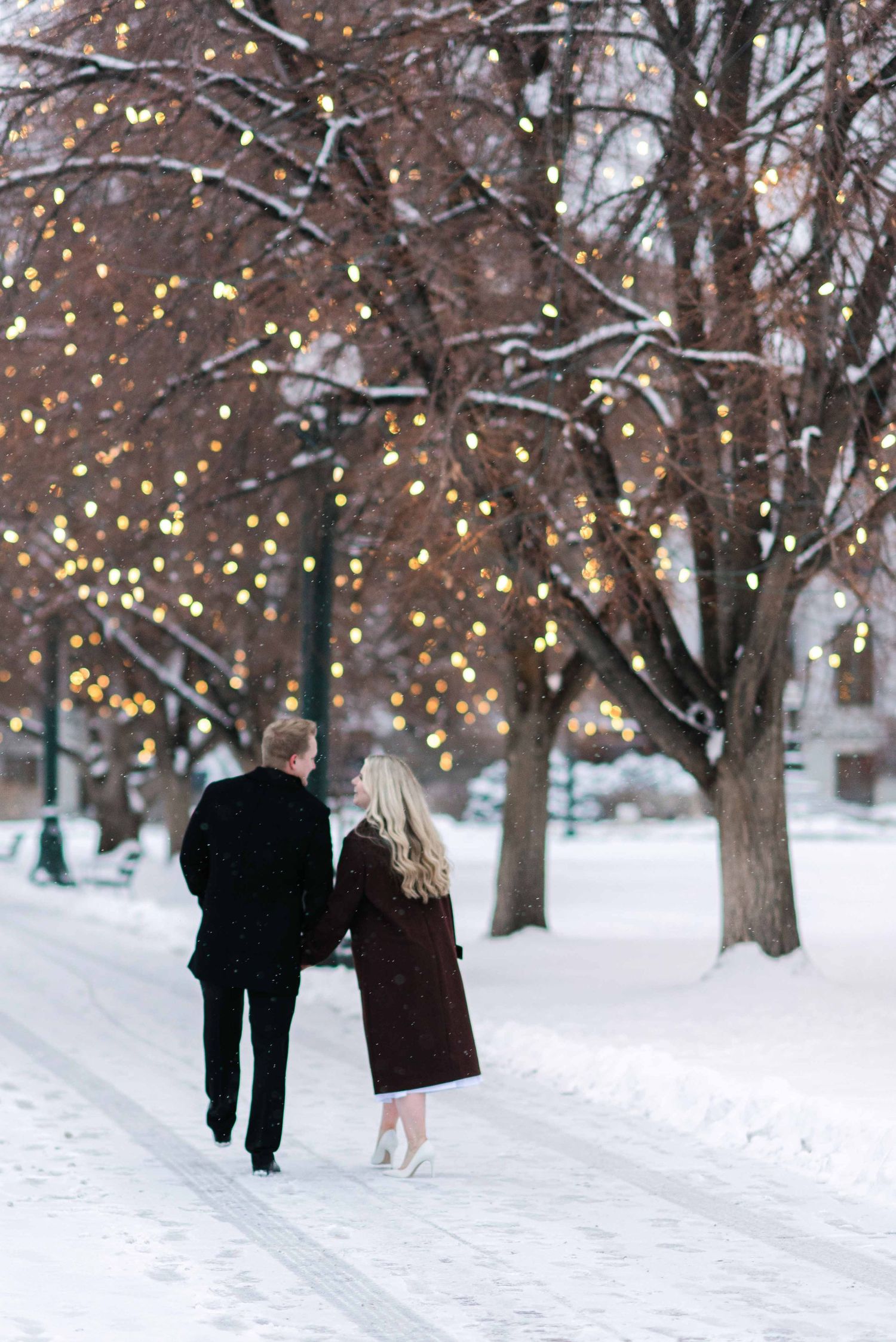A couple walks along a snow-covered path beneath trees with twinkling lights.