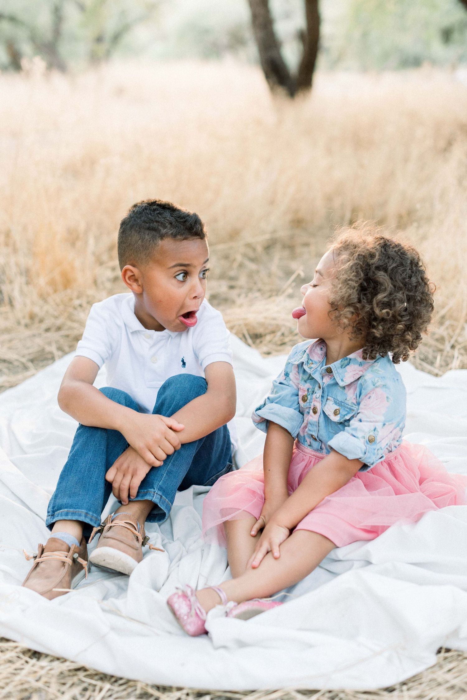 Two children sit on a white blanket making silly expressions and laughing together.