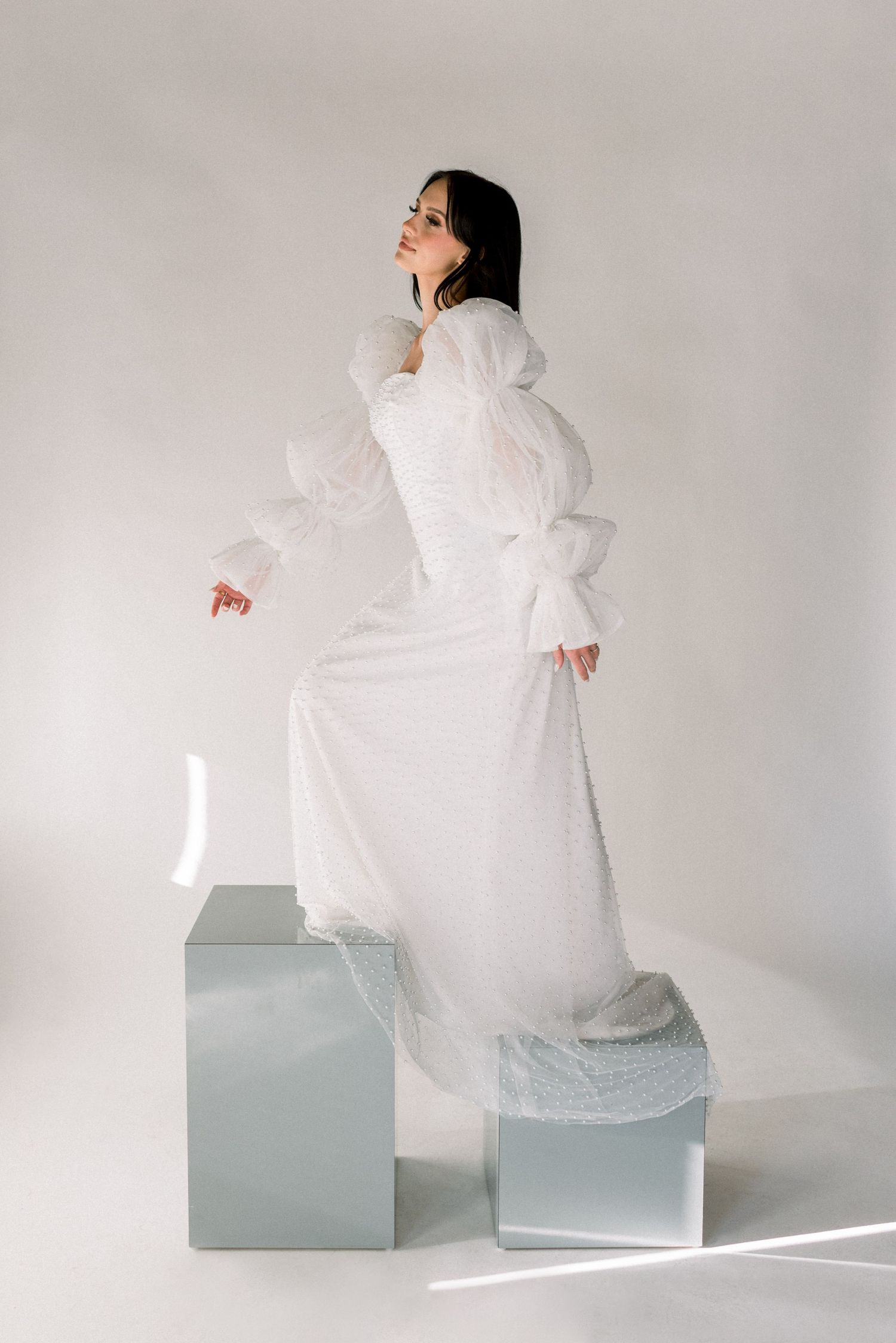Model in textured white gown standing gracefully on geometric blocks in minimalist studio setting.