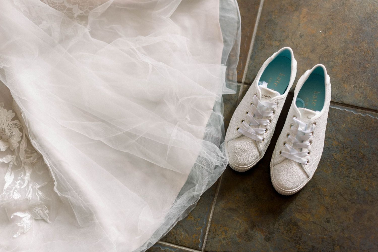 White bridal sneakers rest beside the hem of a flowing tulle wedding dress on stone floor.