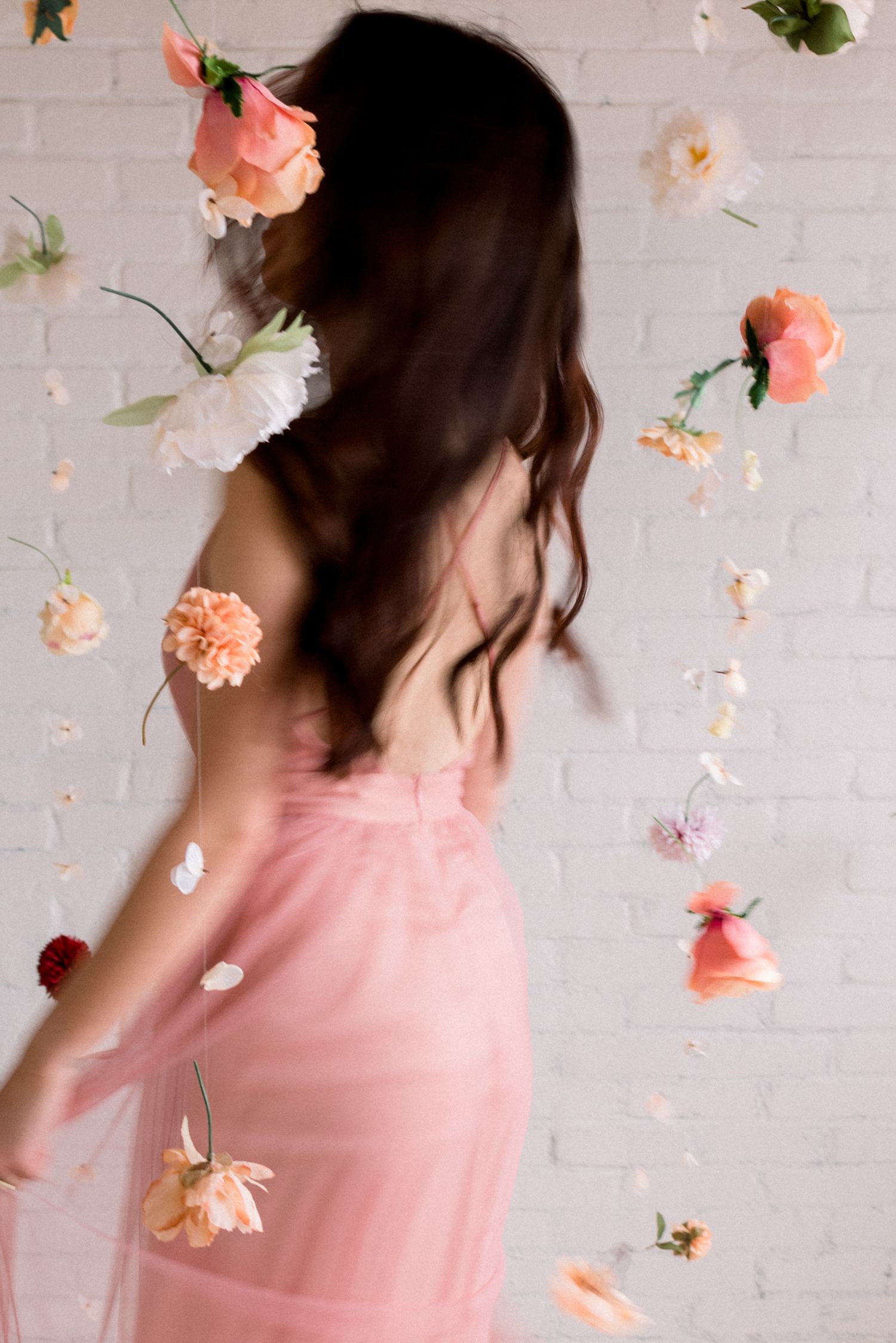 Artistic portrait with floating flowers and pink dress against white brick wall.