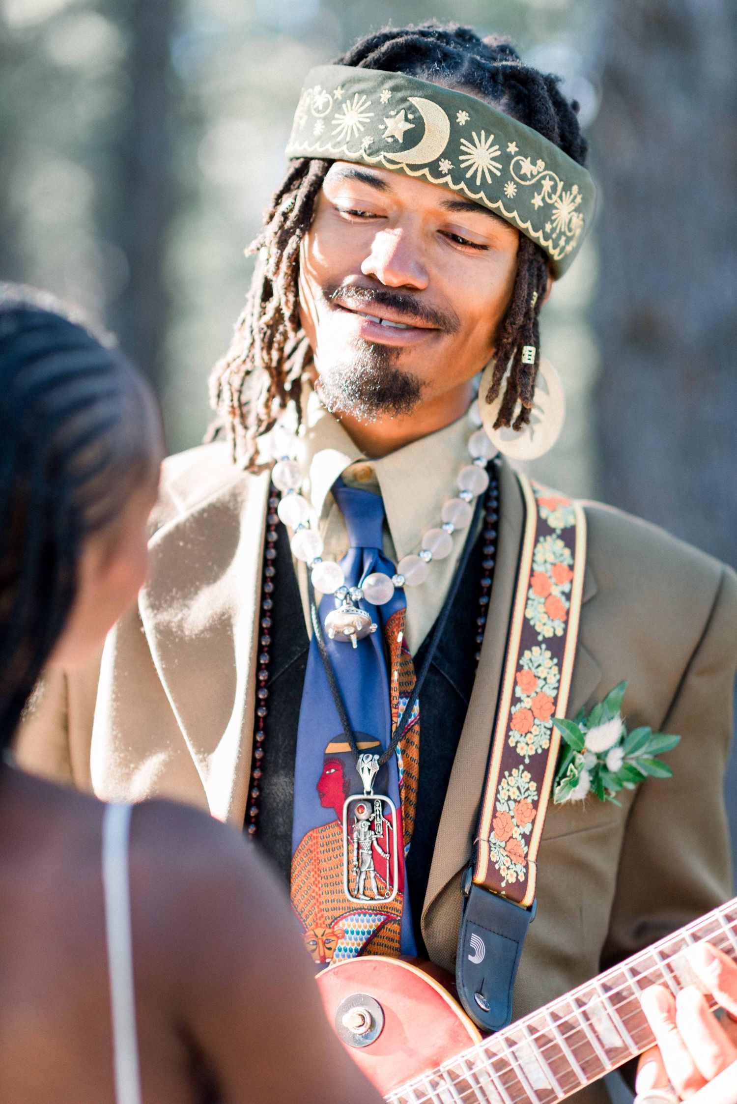 A wedding portrait showing the groom in a headband and suit holding a guitar while smiling.