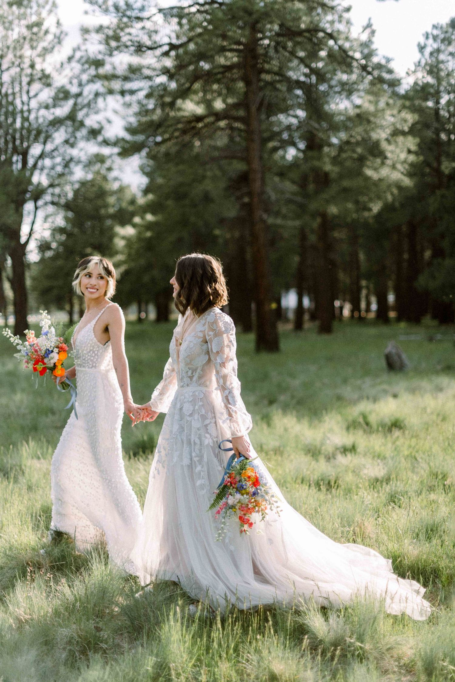 Two brides in flowing white gowns walk hand in hand through a sunlit forest meadow.
