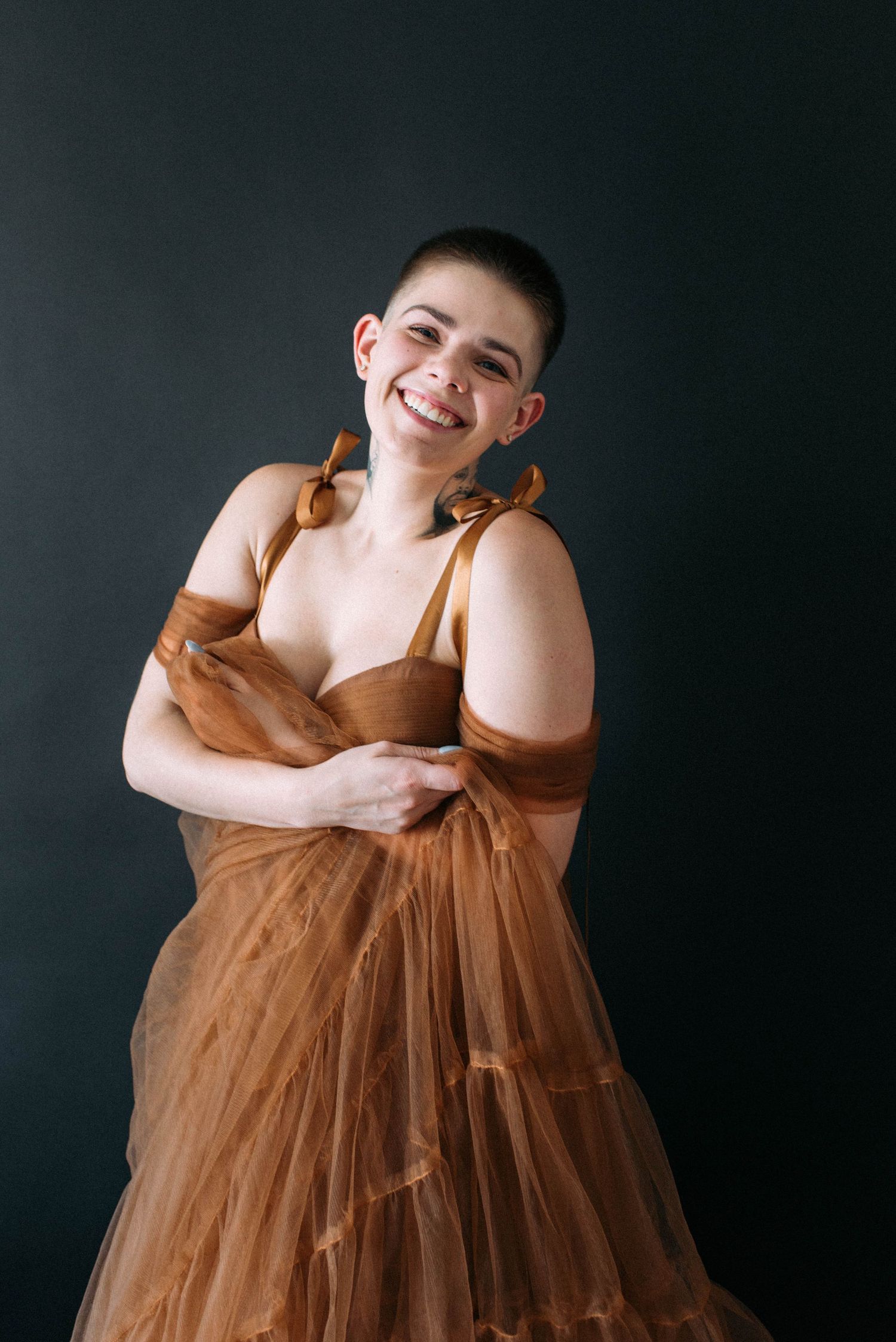 Person in a flowing brown tulle dress poses playfully against a dark backdrop.