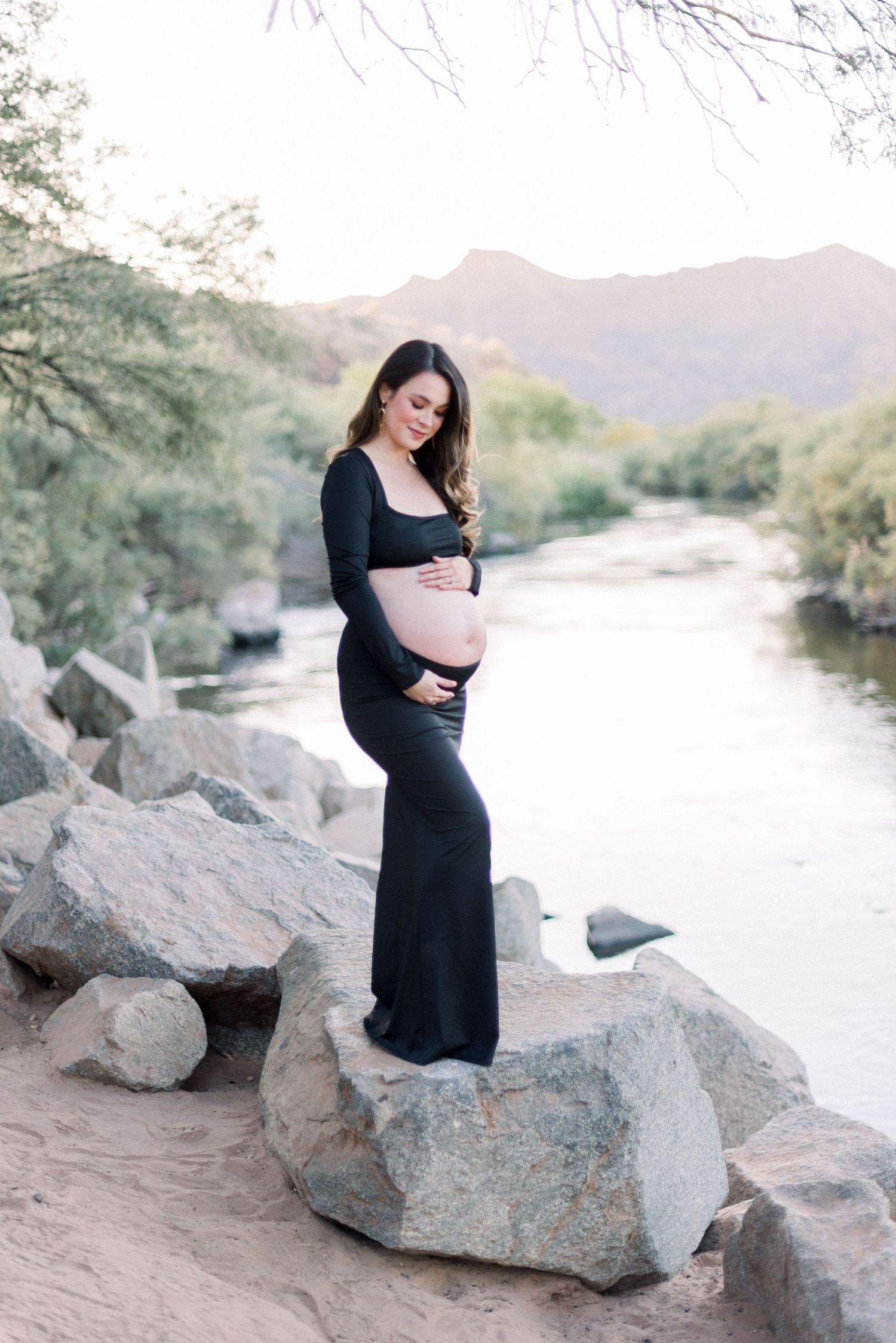 A woman in a long black dress poses gracefully on desert rocks near a winding river at sunset.