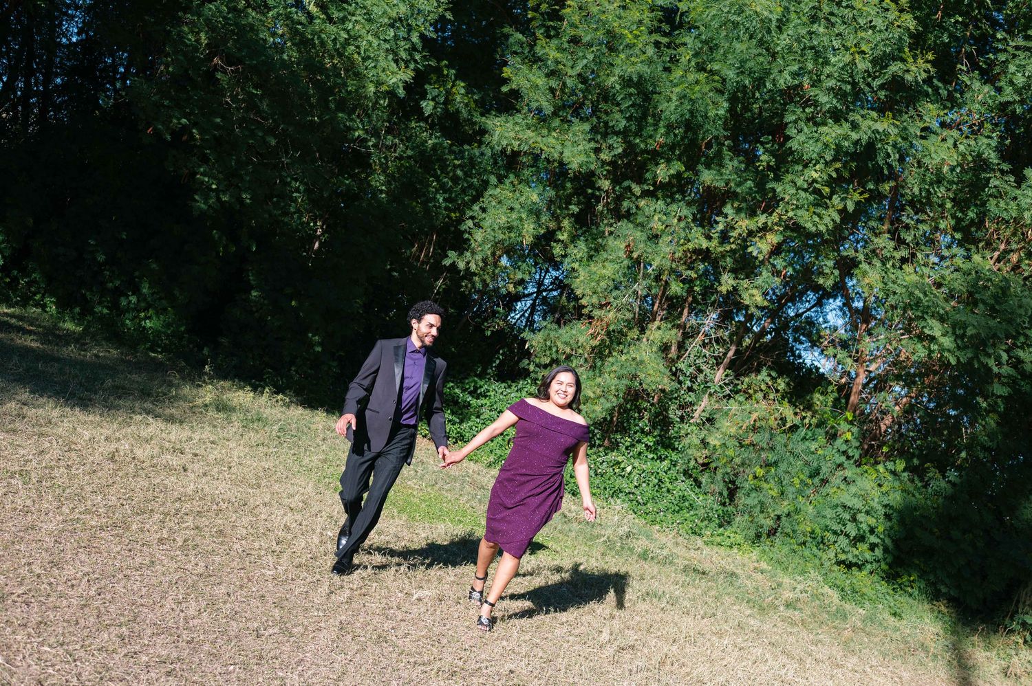 Two people in formal attire hold hands while walking together on a dirt path surrounded by green foliage.