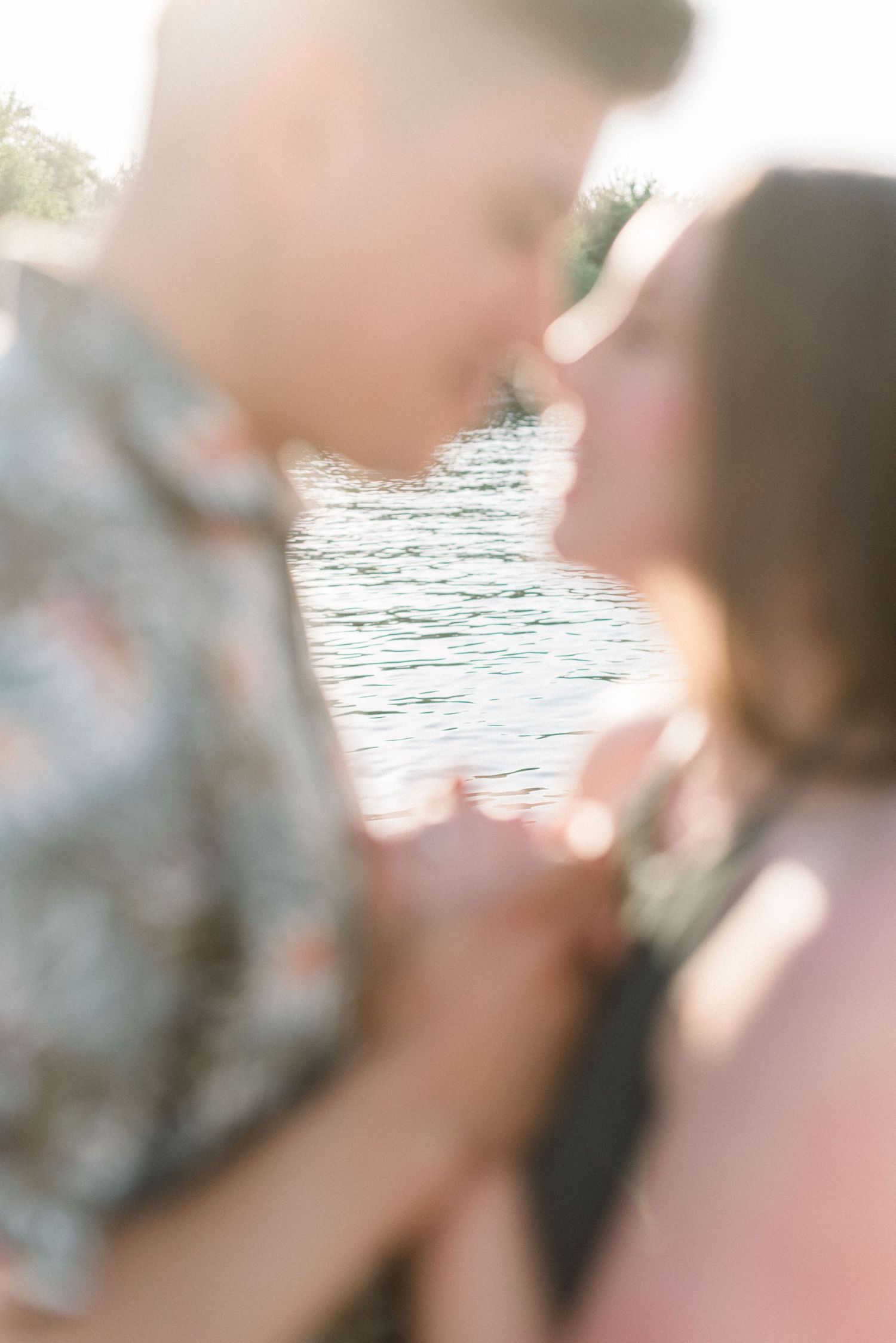 A couple shares a tender kiss while standing near a waterfront with sunlight creating a romantic glow.