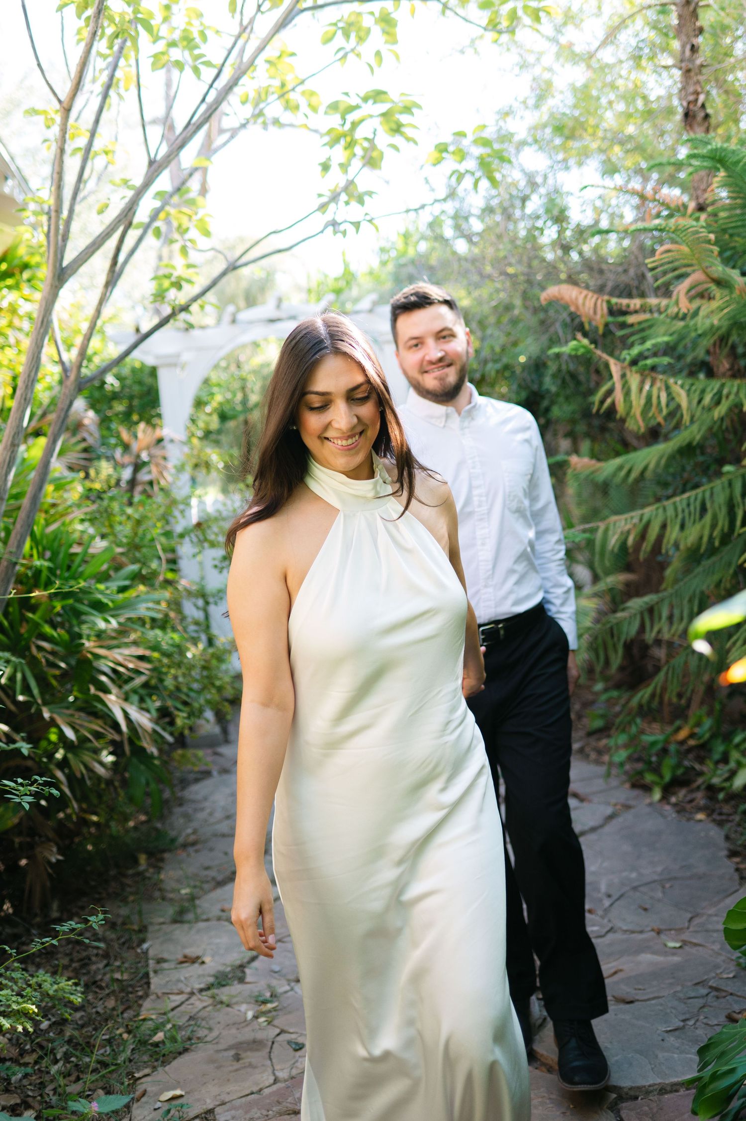 A couple shares a playful dance moment on a garden path surrounded by tropical plants.