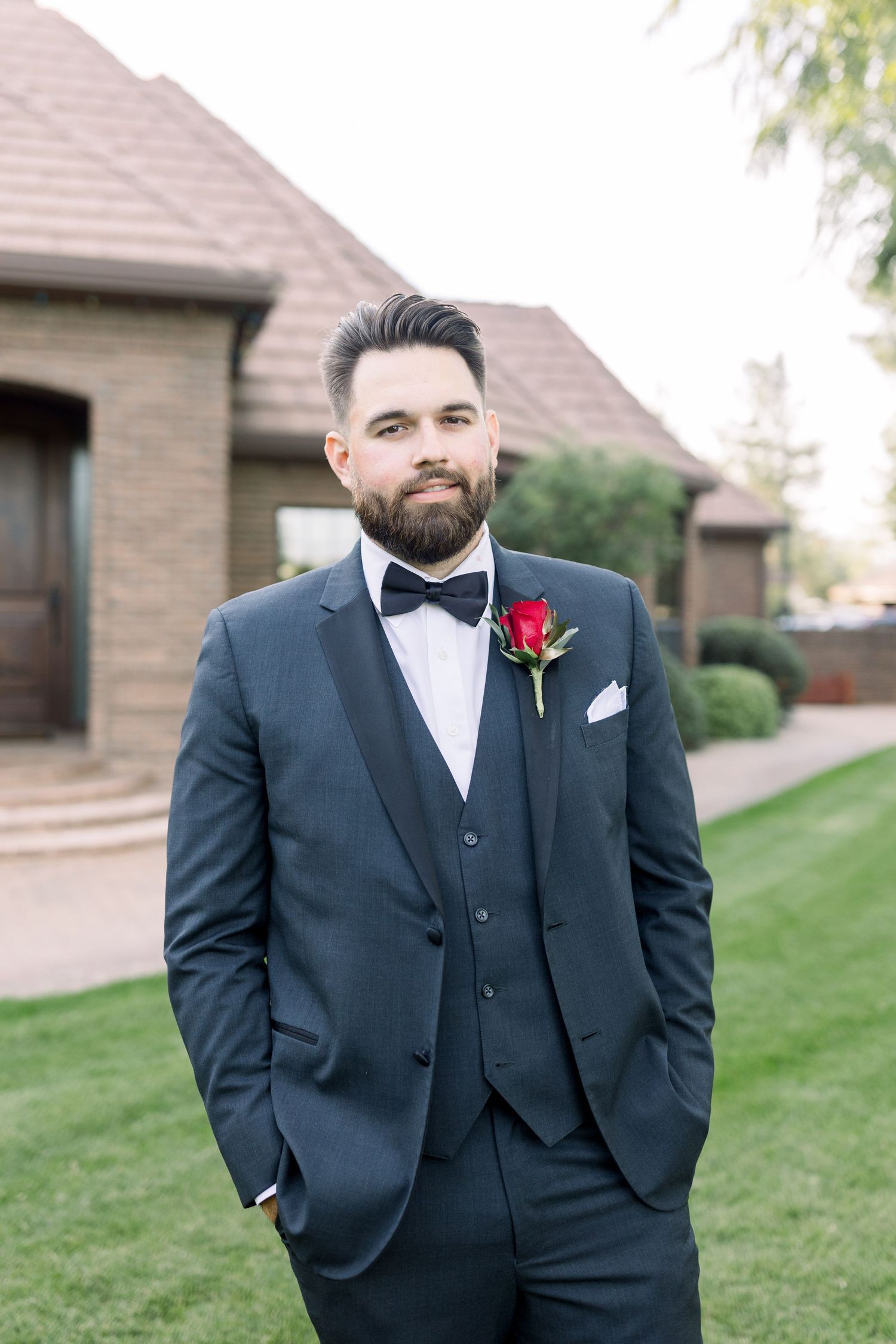 Groom in navy three-piece suit with bow tie and red boutonniere poses on lawn.