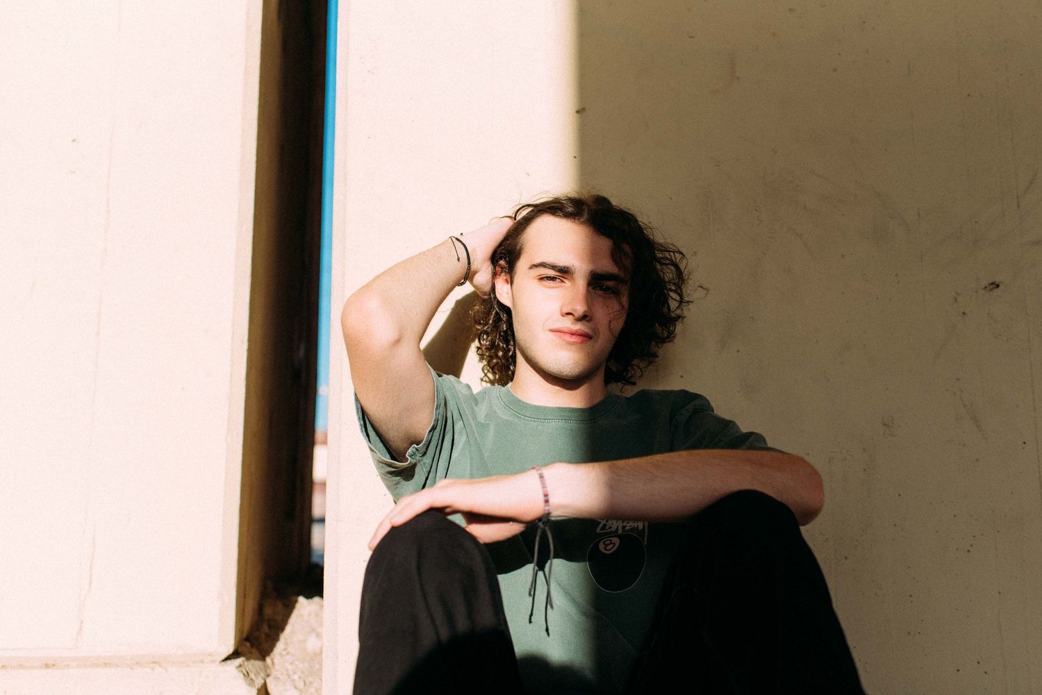 Person with curly hair sitting against wall in casual green shirt and black pants.