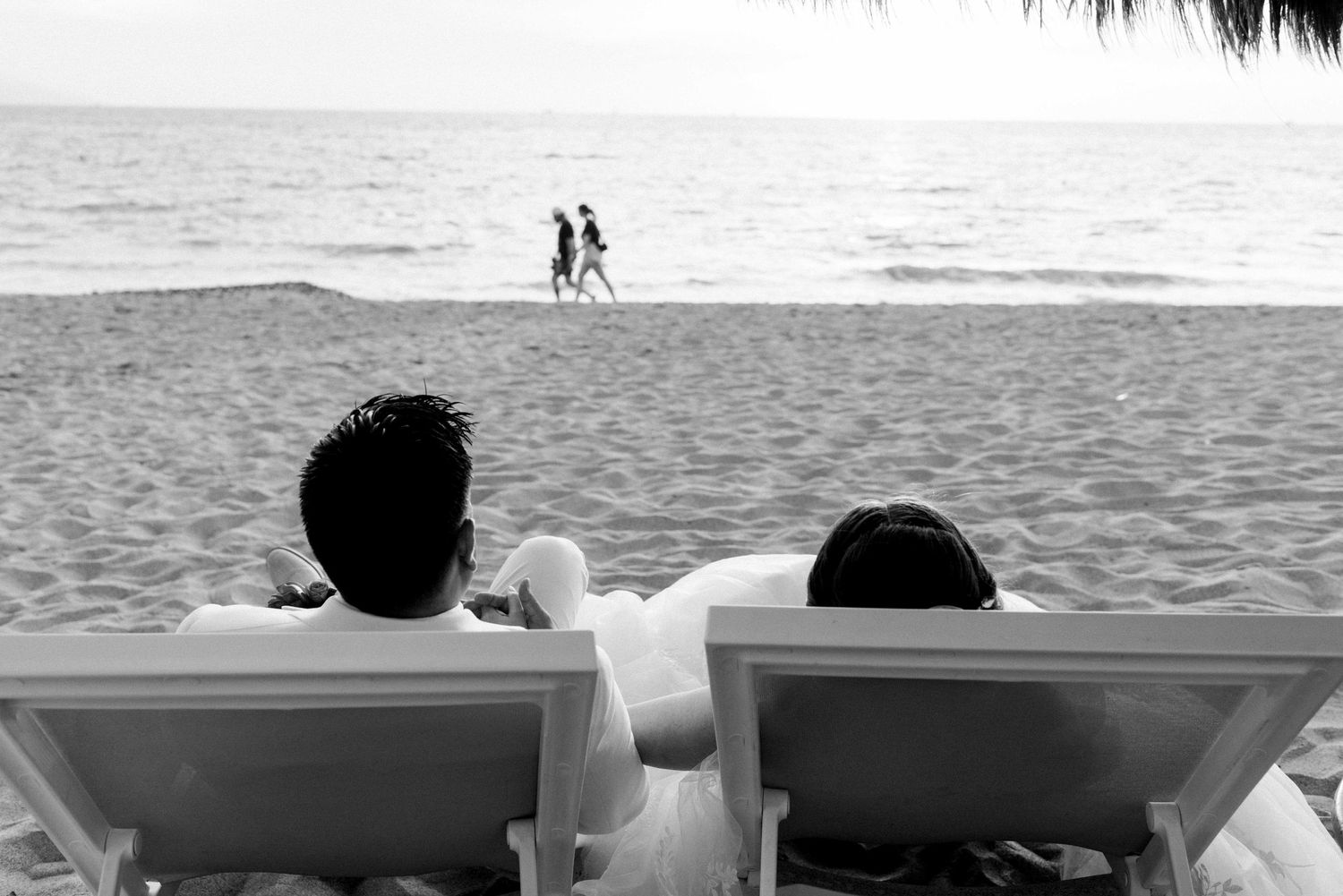 Two people relax in beach chairs watching distant figures walk along the shoreline.
