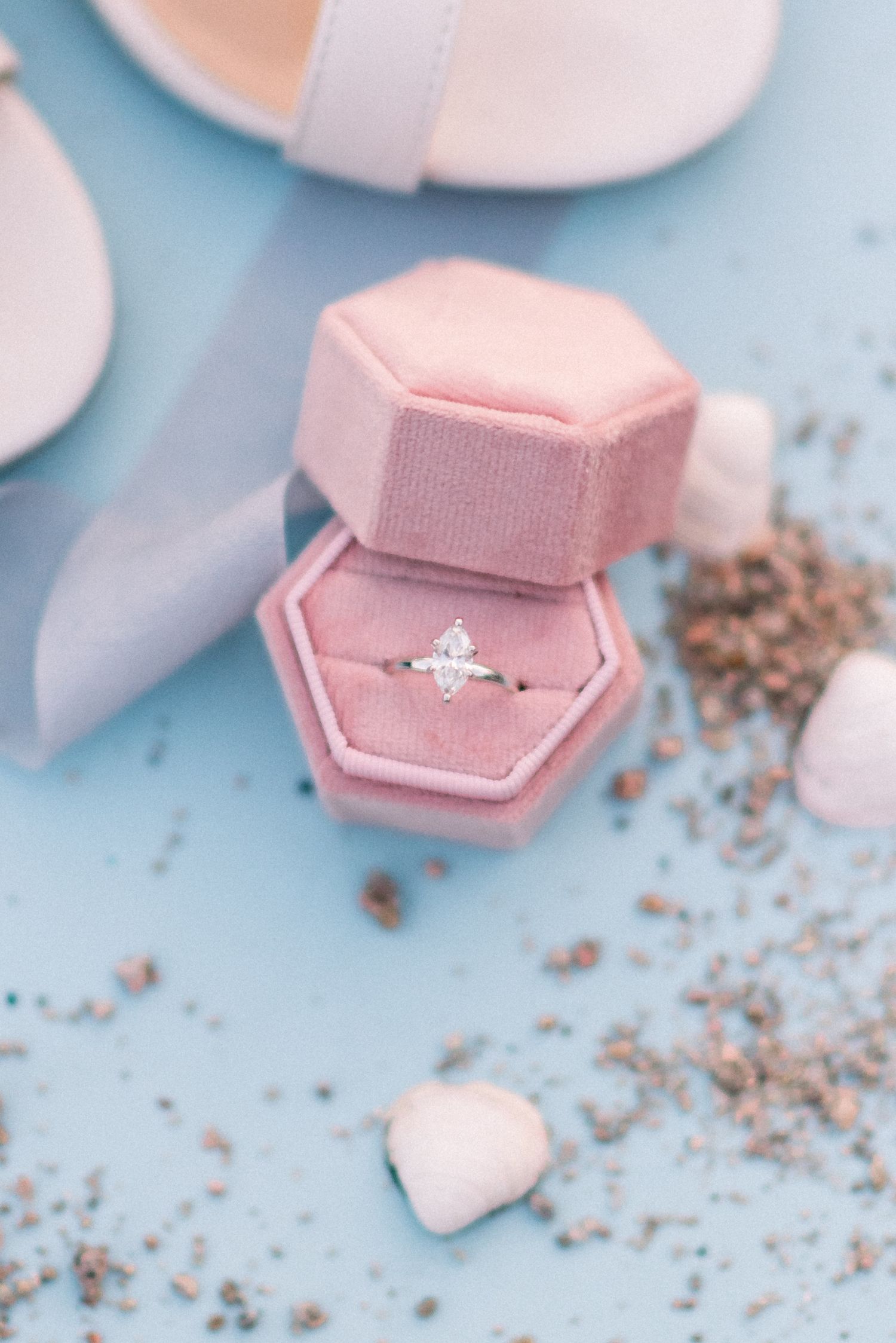 Delicate engagement ring in pink velvet hexagonal box with scattered rose petals on blue background.