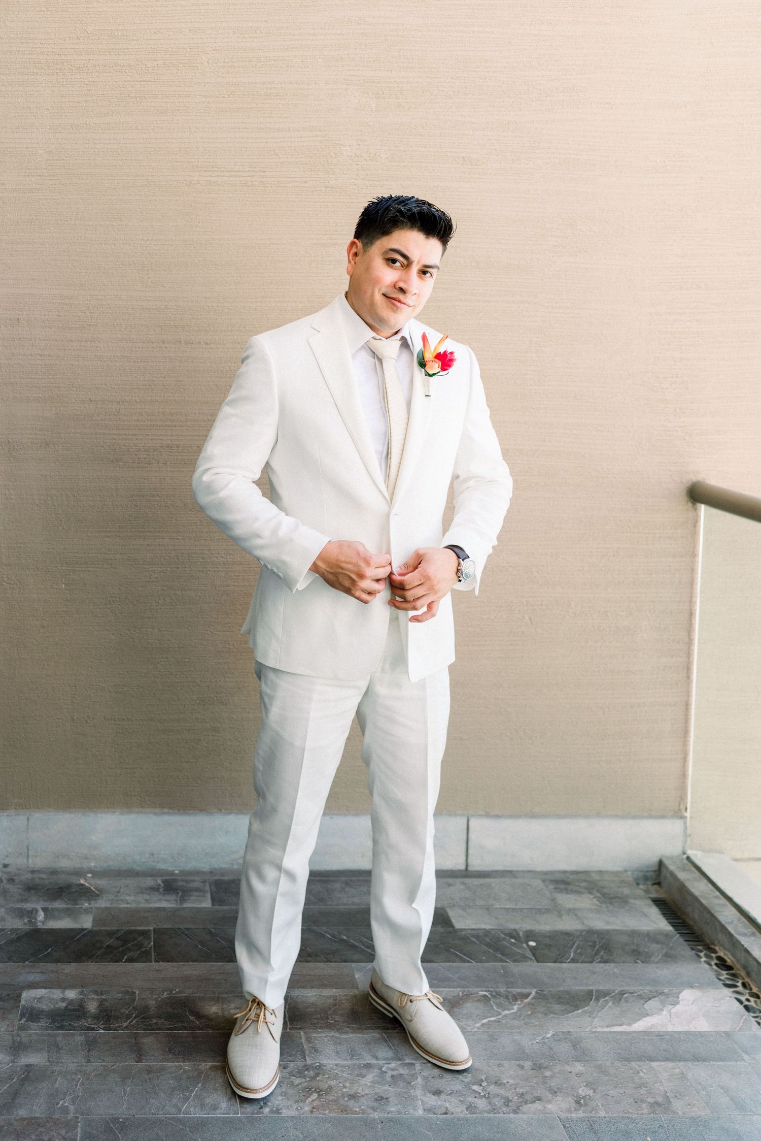 Full-length portrait of groom in white suit with red boutonniere against neutral wall.