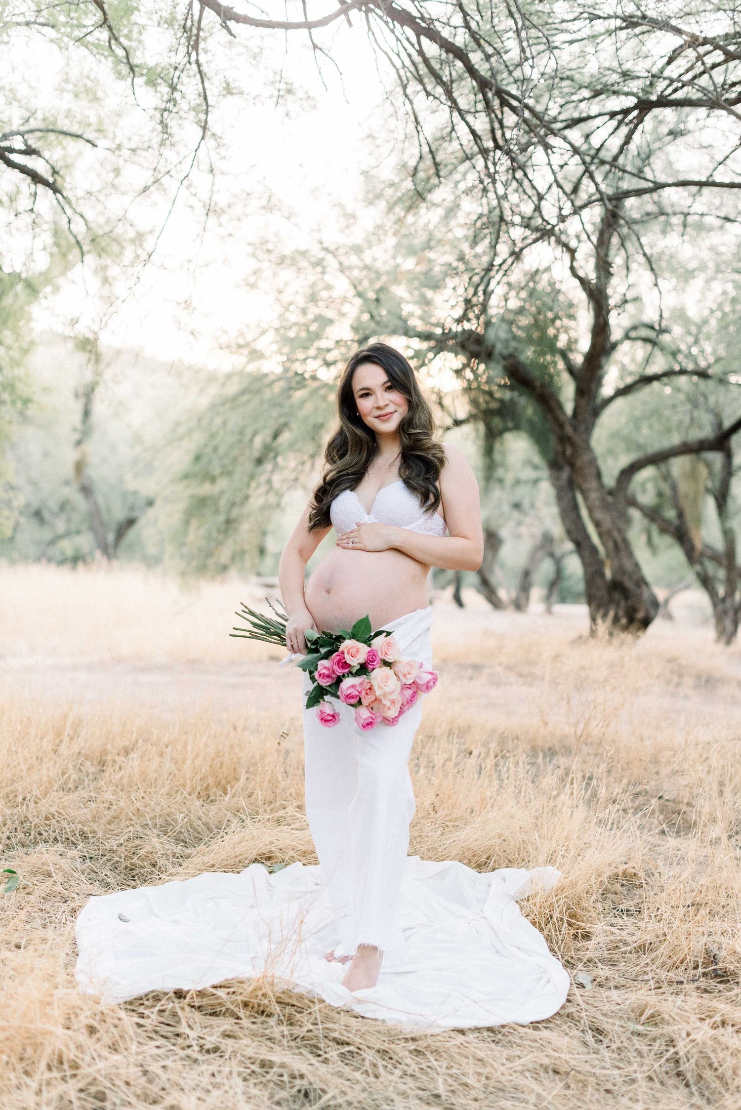 Outdoor maternity portrait with pink bouquet and flowing white dress in golden field.