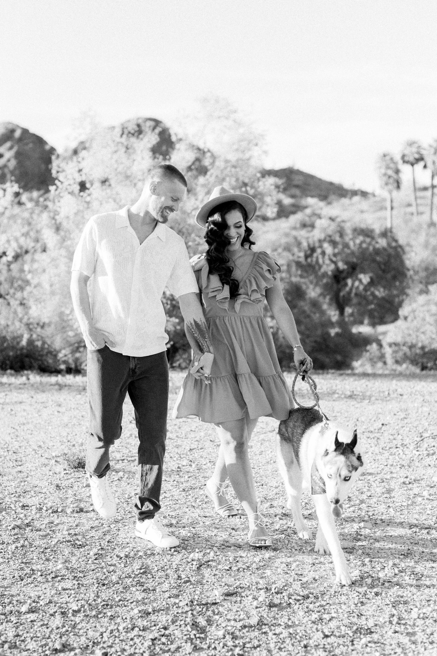 Black and white photo of couple walking with dog against mountain backdrop.