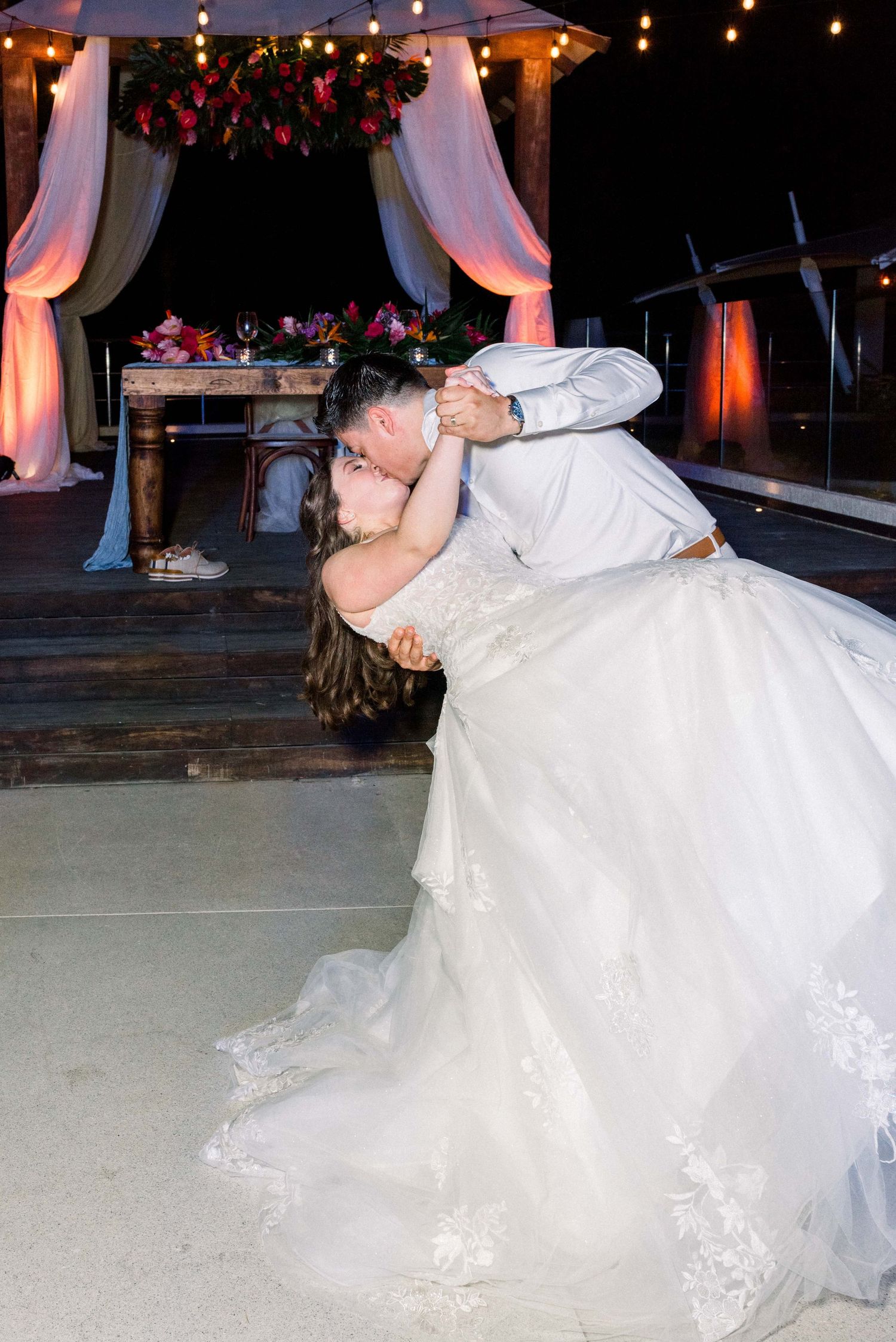 Romantic wedding dip kiss under draped fabric and string lights at outdoor reception.