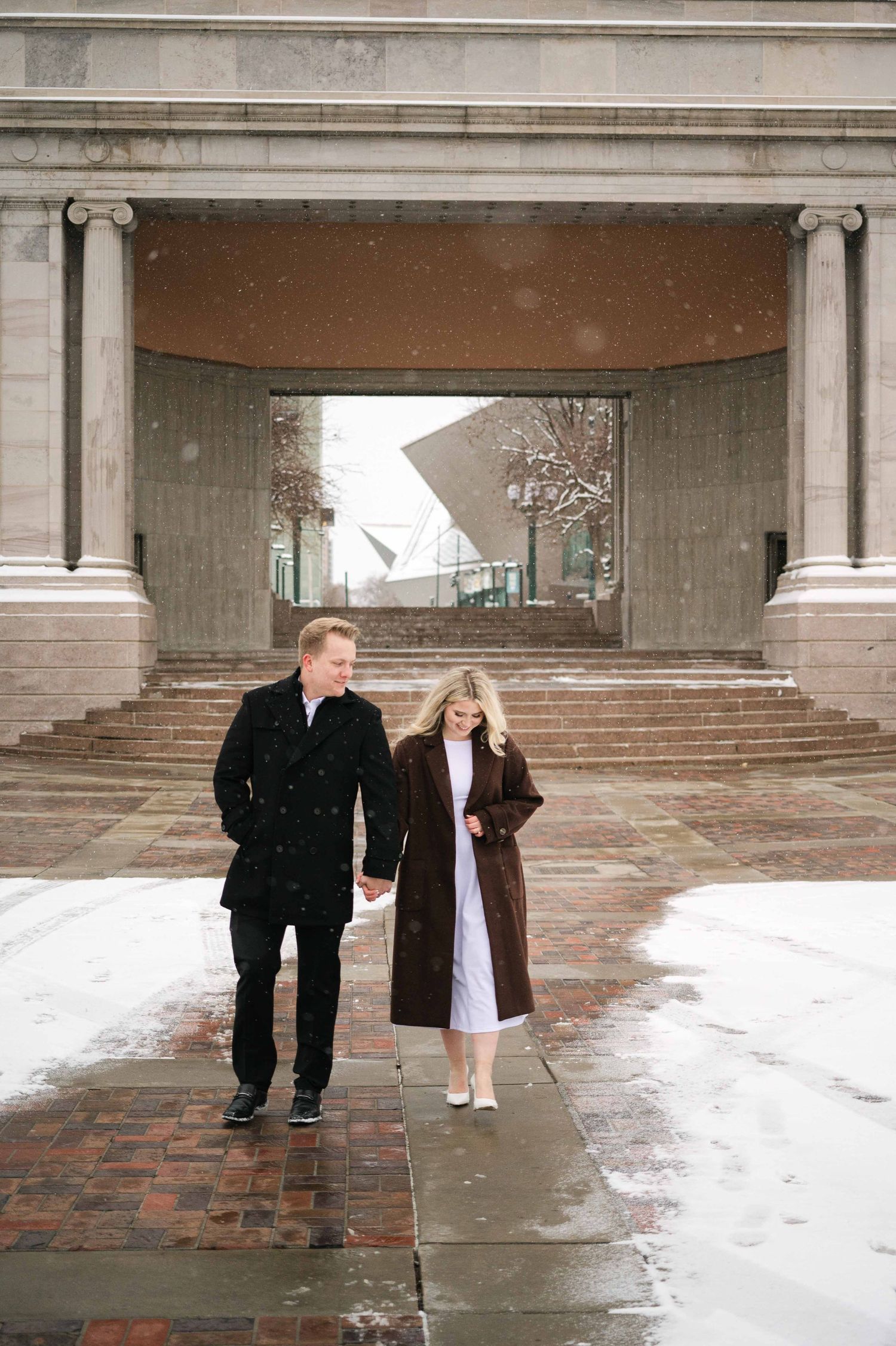 A couple walks hand in hand through a snowy colonnade entrance of a classical building.
