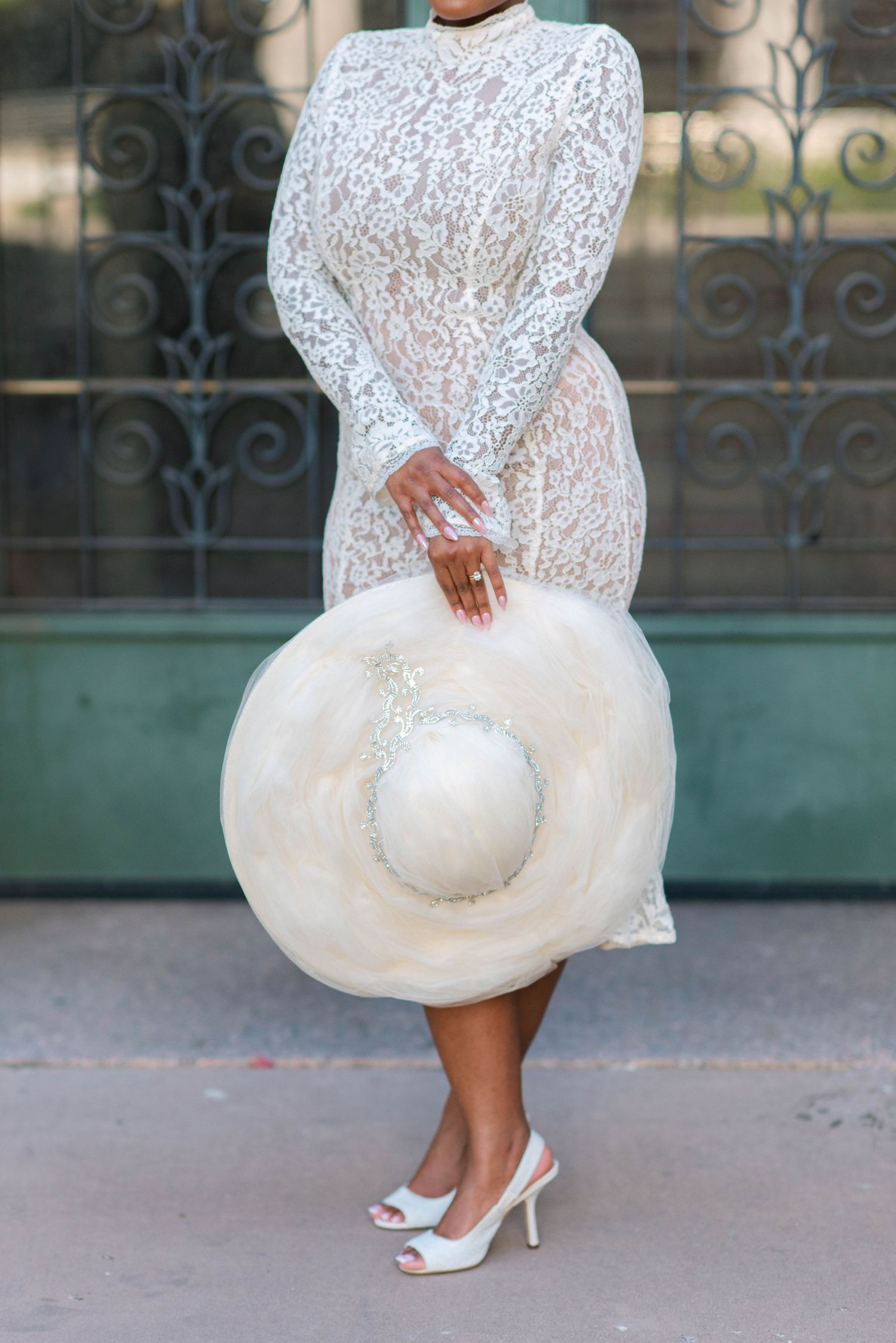 A fitted white lace dress with long sleeves is styled with a dramatic wide-brimmed hat and peep-toe heels.
