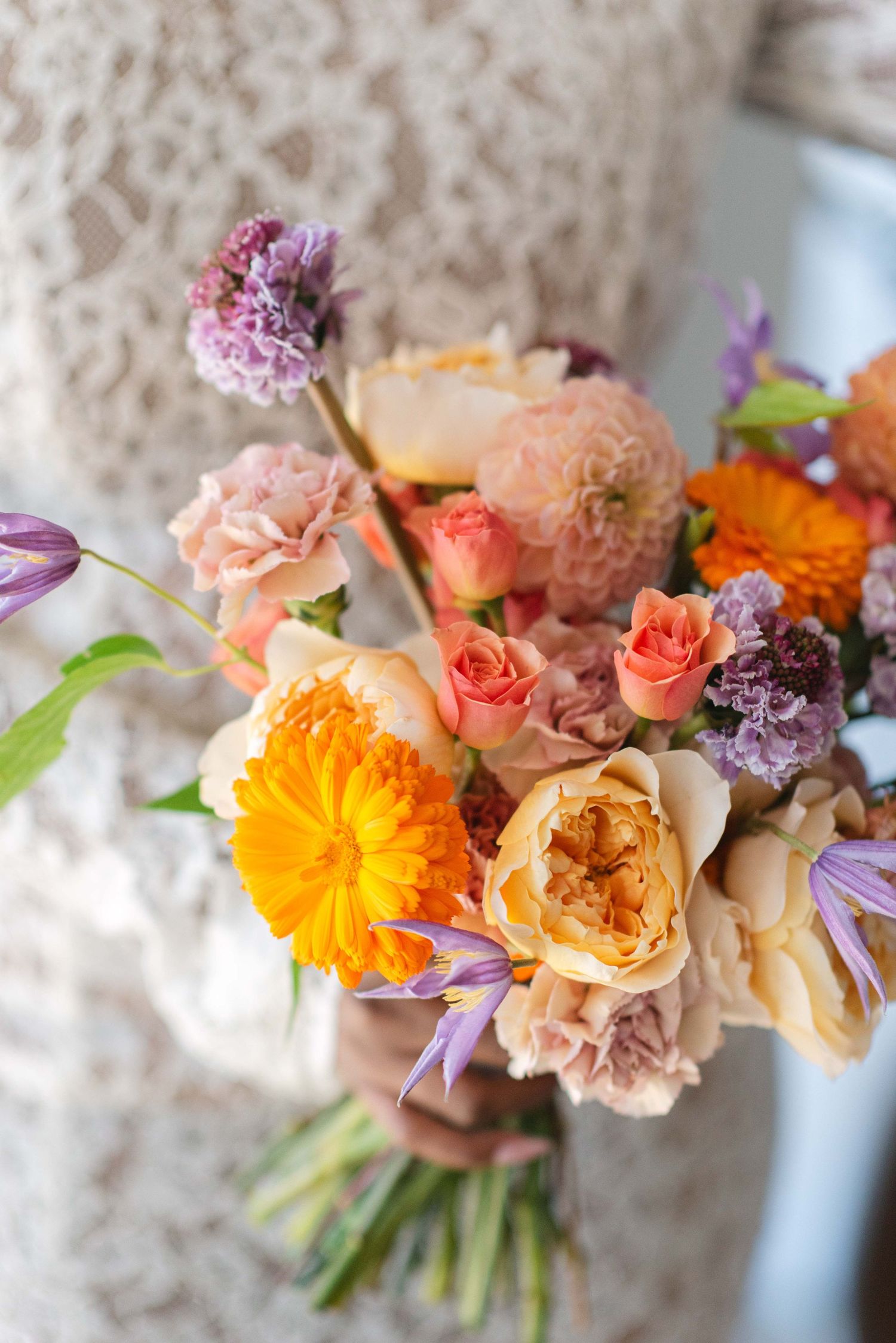 Vibrant wedding bouquet with orange, pink, and purple flowers against lace background.