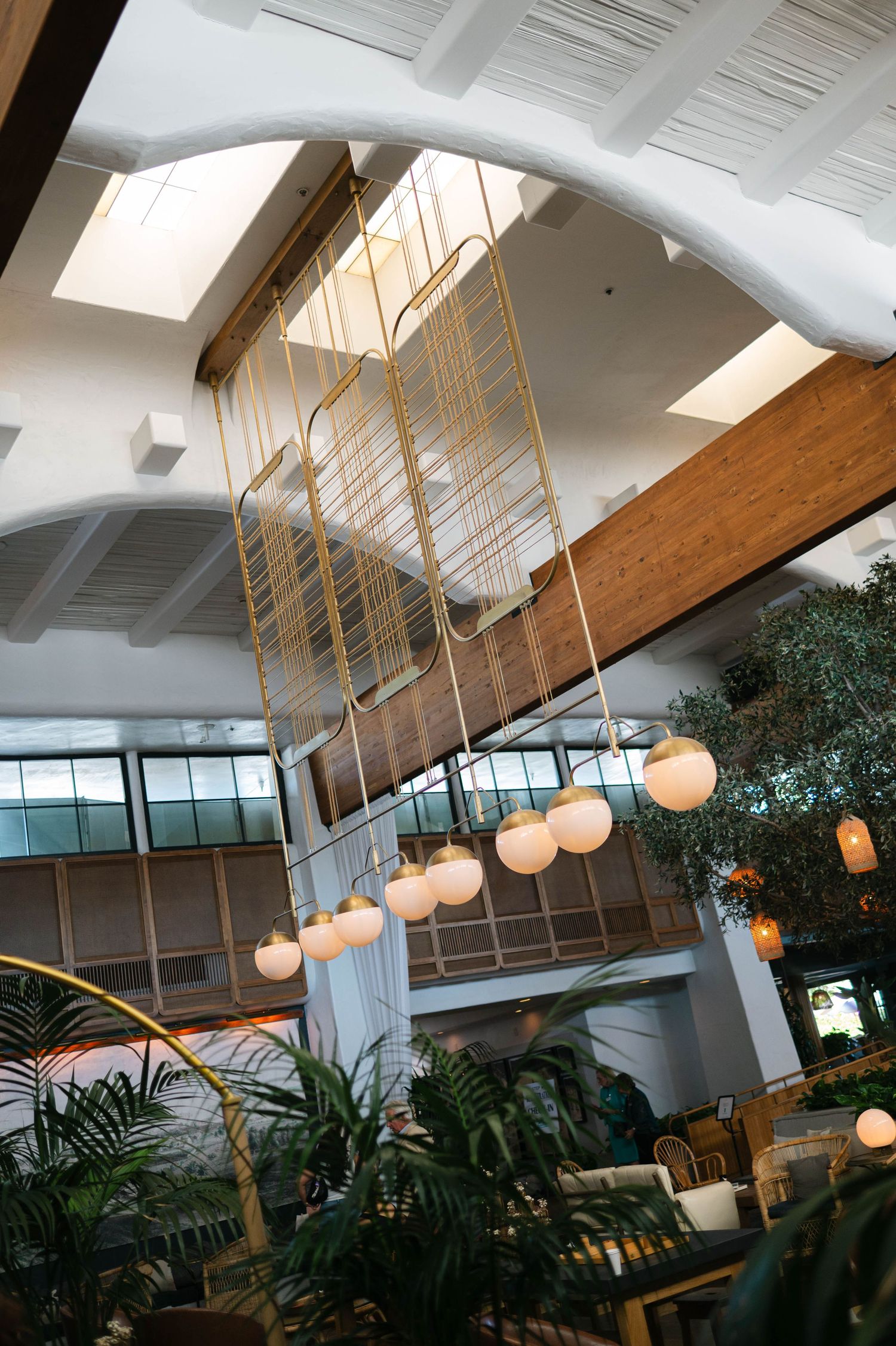 Modern office interior with hanging pendant lights, wooden beams, and lush indoor plants beneath a skylit ceiling.