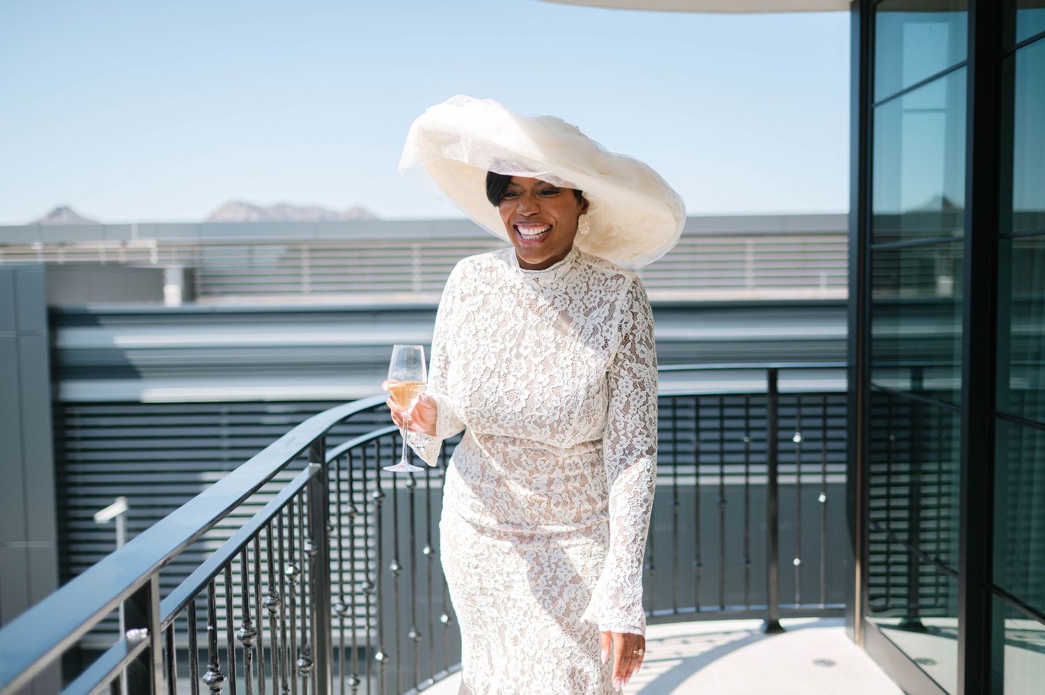 A bride in a white lace dress and wide-brimmed hat poses on modern glass building steps.
